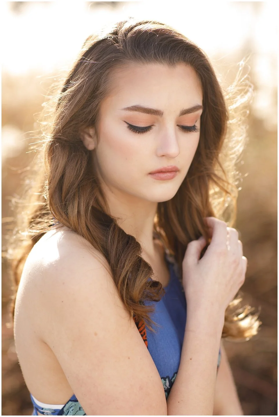 A young woman with long, wavy brown hair and closed eyes, dressed in a sleeveless blue top, standing outdoors with sunlight backlighting her hair.