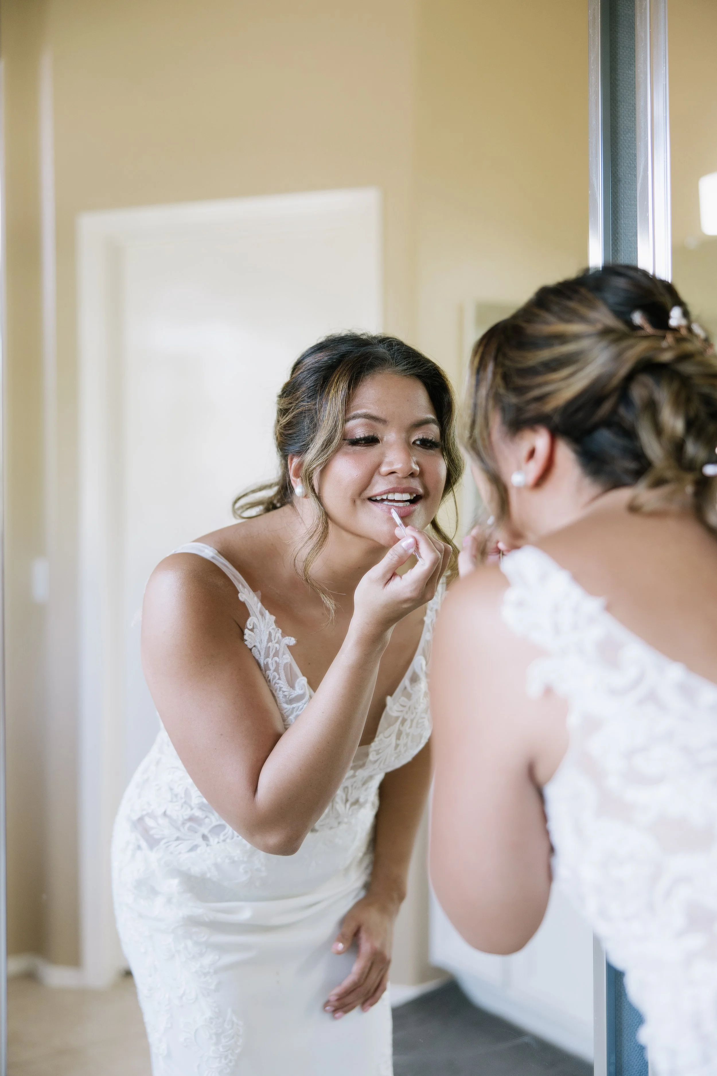 Woman in wedding dress applying lipstick in front of mirror.