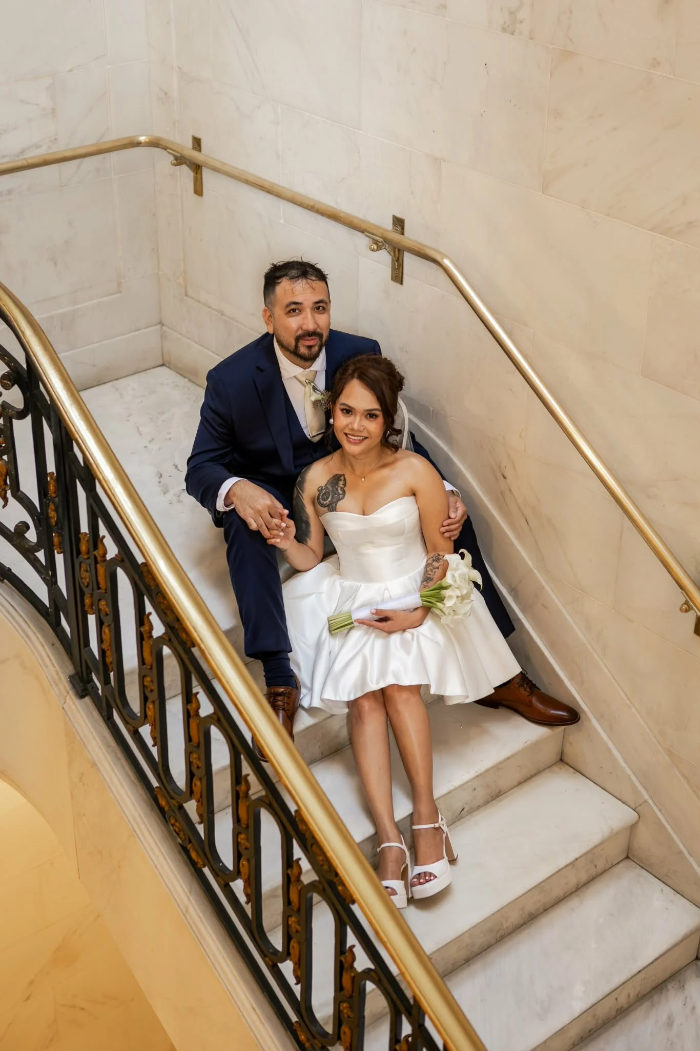 A newlywed couple sitting on marble stairs inside a building with cream-colored walls. The man is in a navy suit with a beige tie, and the woman in a strapless white wedding dress holding a bouquet of white flowers. They are both smiling and looking 