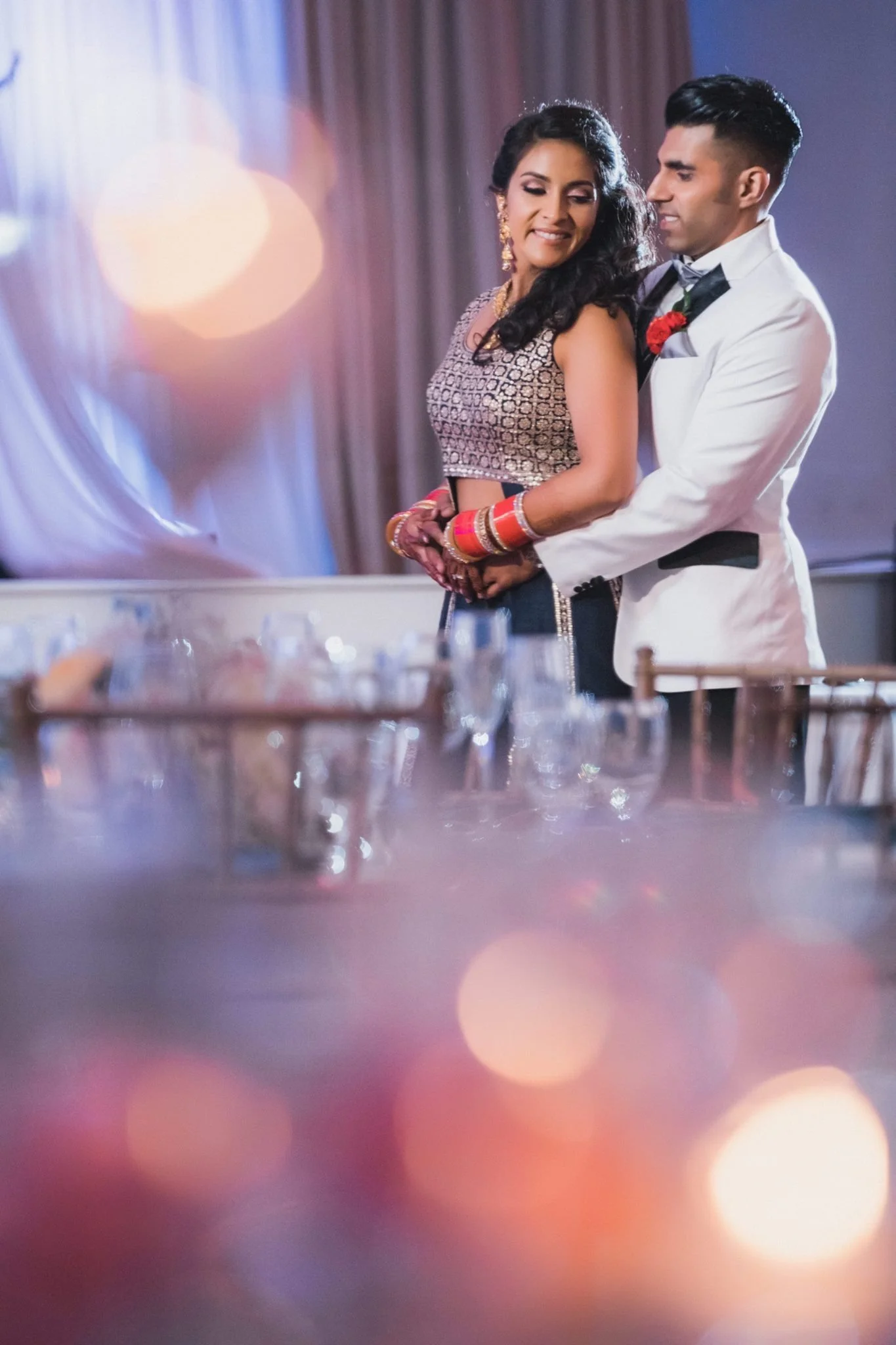 A couple dancing at a wedding reception, with the woman wearing traditional Indian jewelry and attire, and the man dressed in a white tuxedo. They are smiling, close together, in an elegant venue with soft lighting.