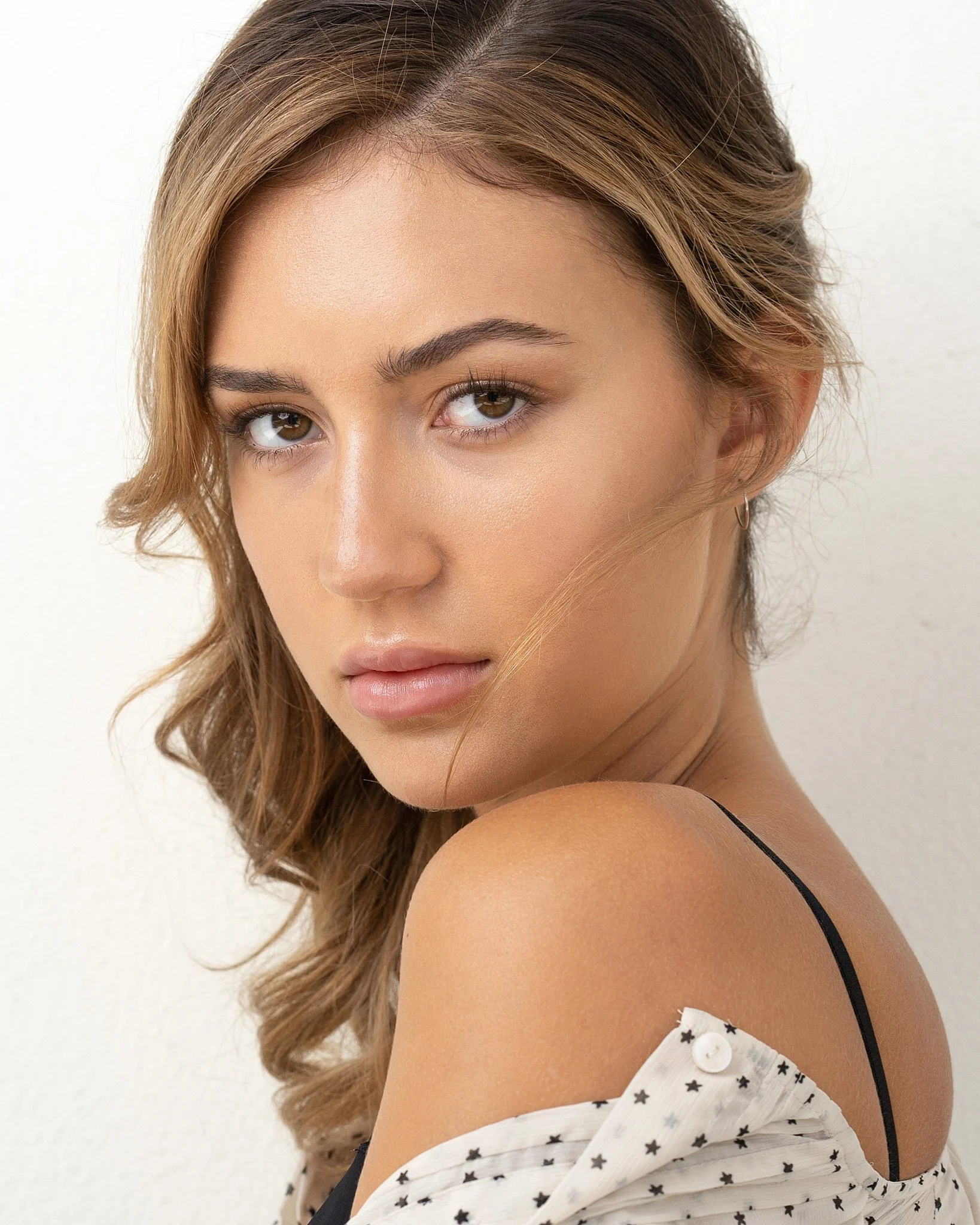 A woman with light brown hair styled in loose curls, wearing a black strap top with a white off-shoulder shirt that has black star patterns and a button detail.