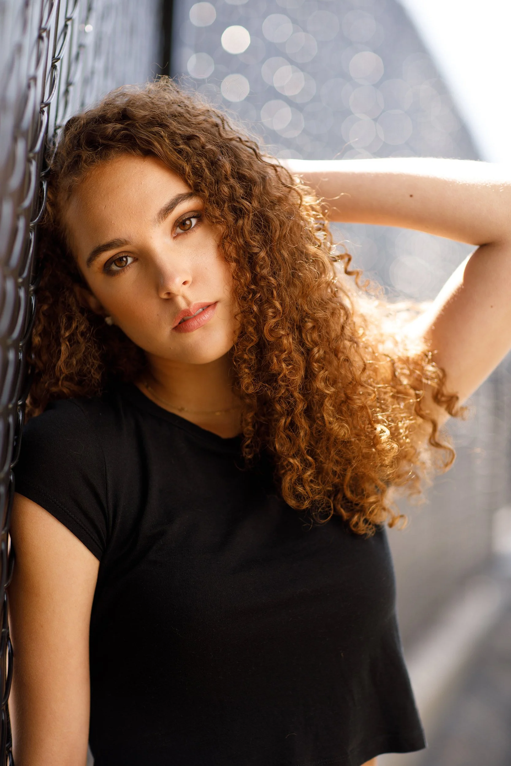 A woman with curly red hair wearing a black shirt, leaning against a chain-link fence with her left arm raised and hand behind her head, looking at the camera with a neutral expression.
