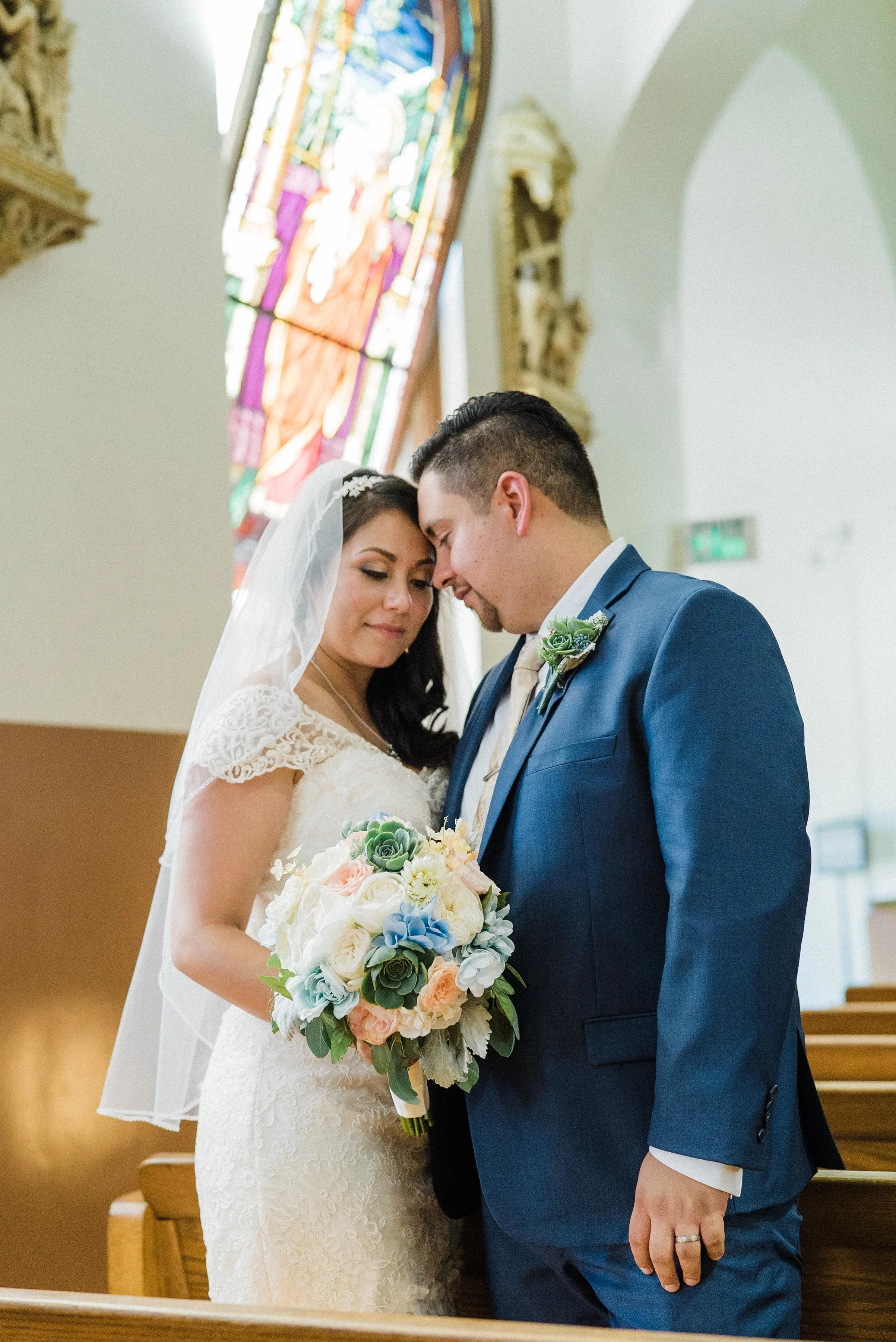 A bride and groom with their foreheads touching, standing close in a church during their wedding ceremony, with a stained glass window in the background.
