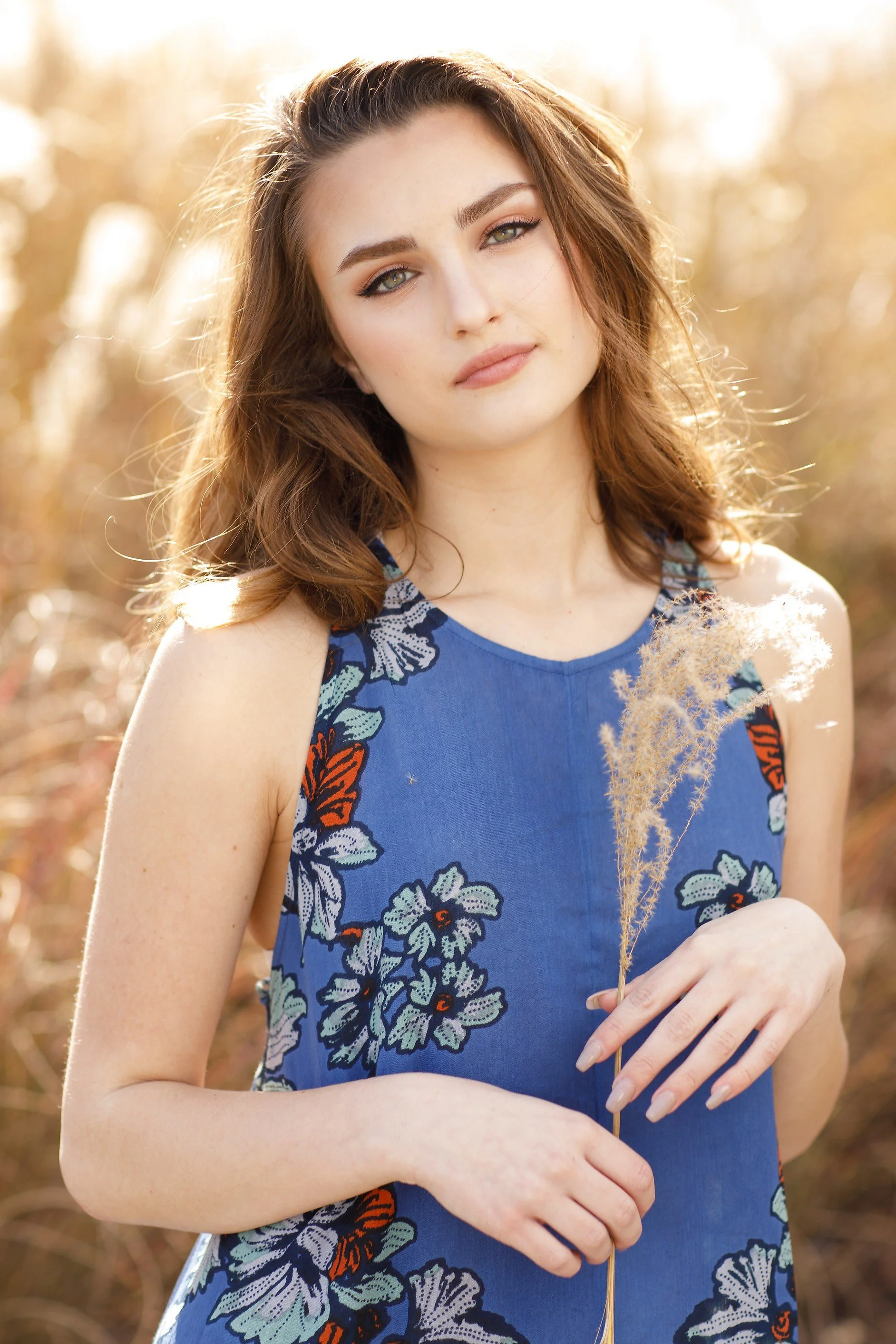 A woman with light skin, brown hair, and blue eyes wearing a blue sleeveless floral dress, holding a dried plant, standing outdoors with a blurred background and sunlight shining behind her.