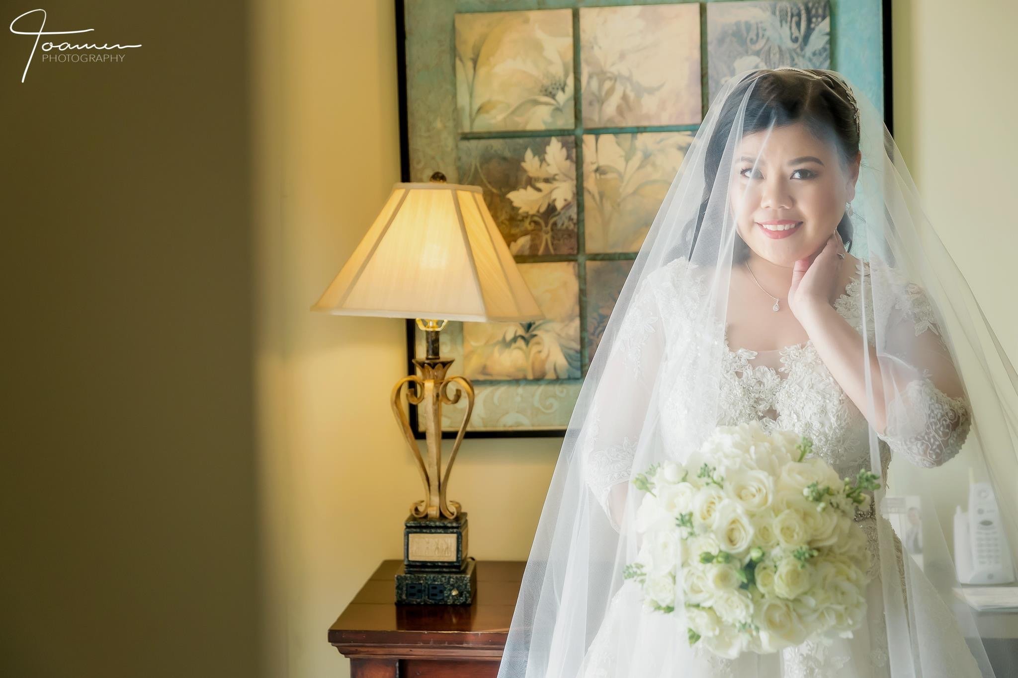 A bride in a white lace wedding dress and veil holds a bouquet of white roses and smiles, standing near a window with a floral painting and a table lamp in the background.