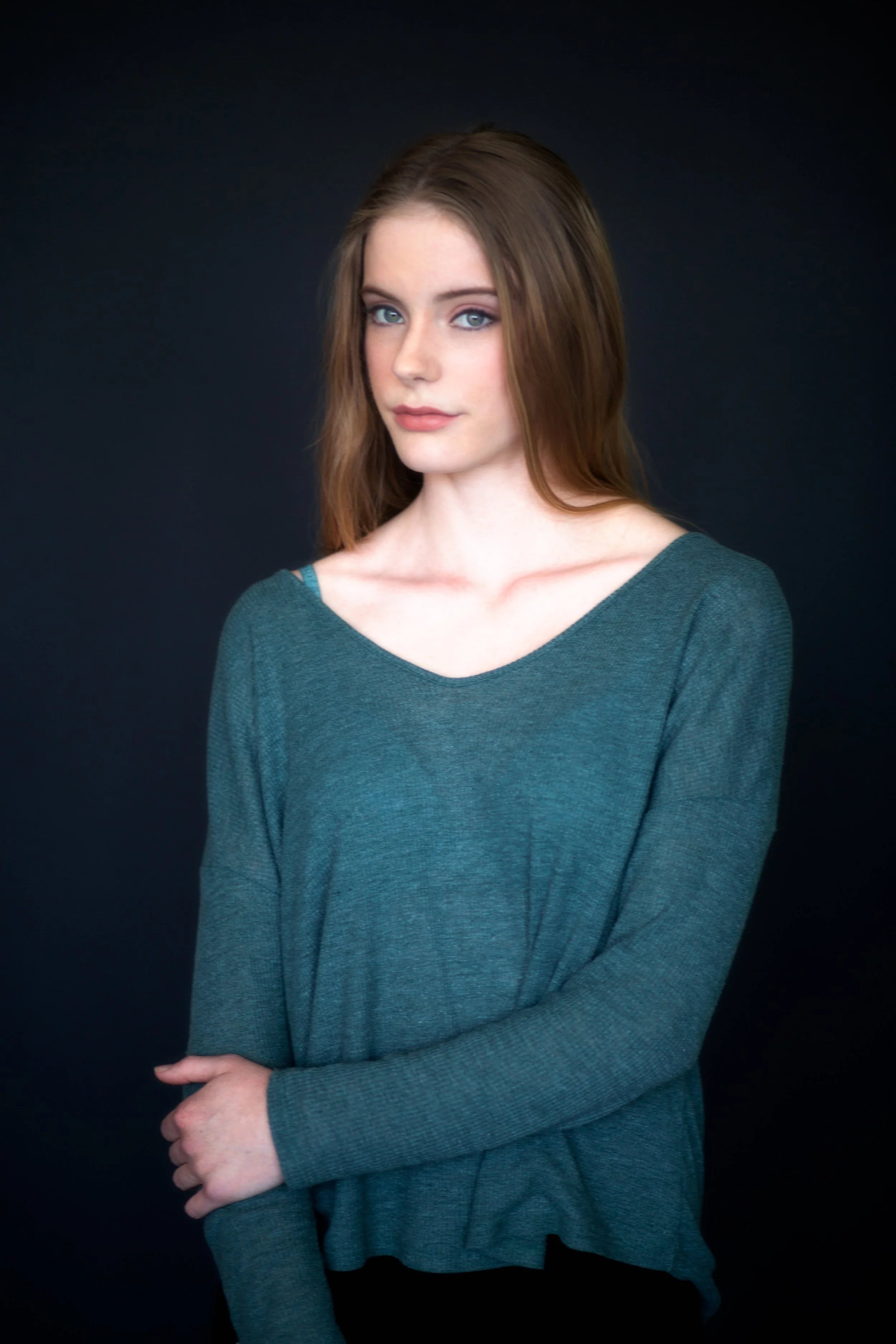 A young woman with long brown hair and blue eyes wearing a teal long-sleeve shirt, standing against a dark background.