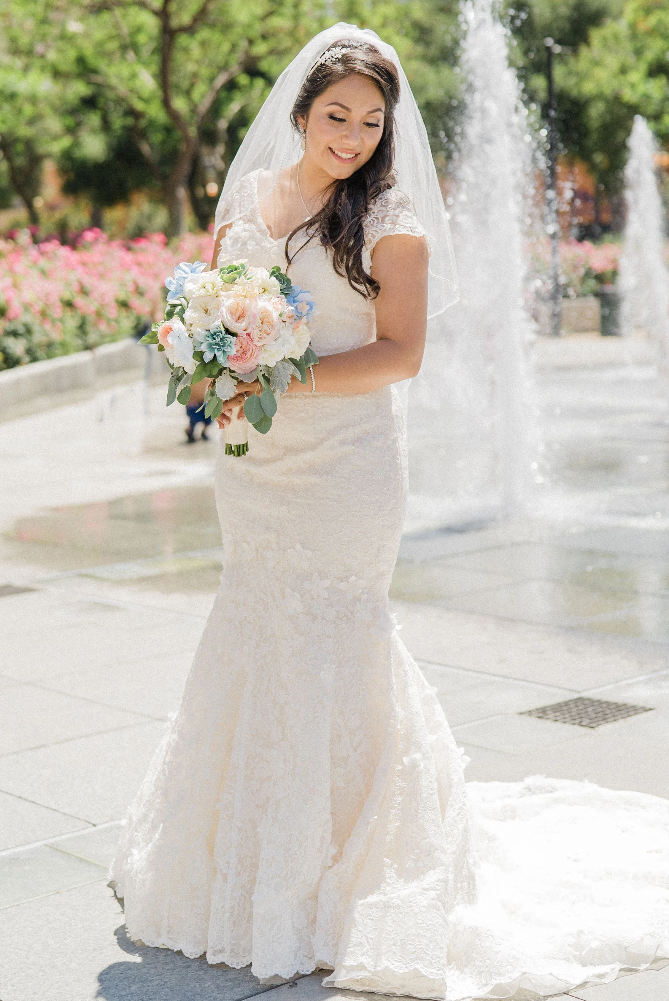 A bride in a white lace wedding dress holding a bouquet of pastel-colored flowers, standing near a fountain in a park with pink flowers and green trees.