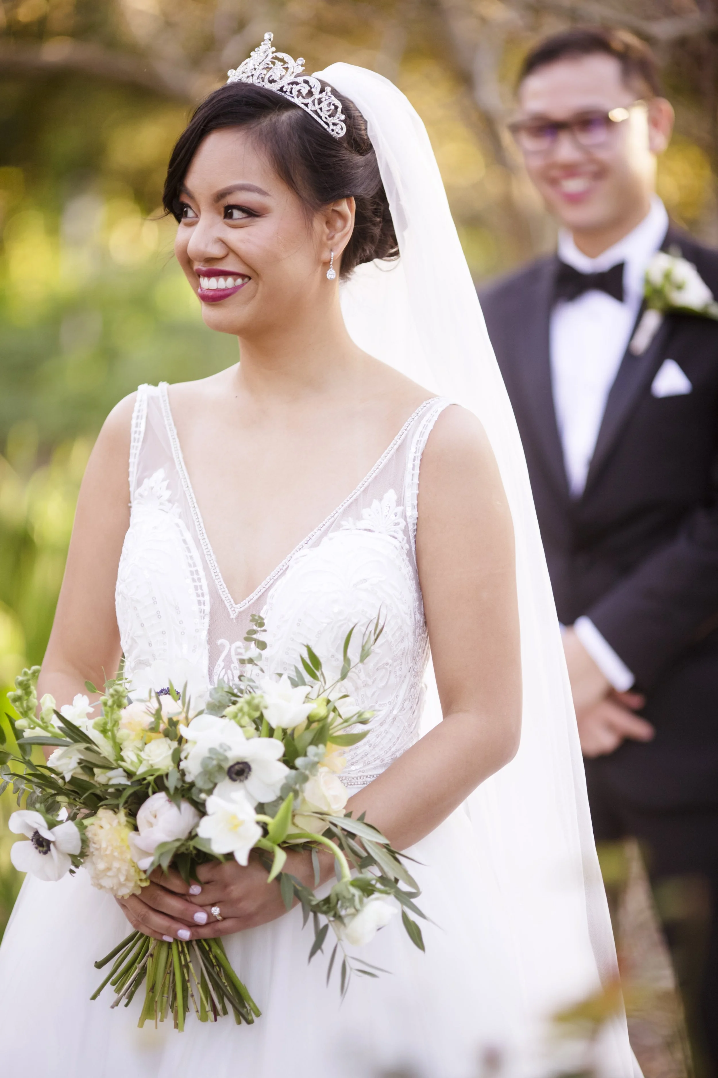 A bride in a white wedding dress and tiara holding a bouquet of flowers, smiling outdoors. A groom in a black tuxedo with a bow tie stands in the background.