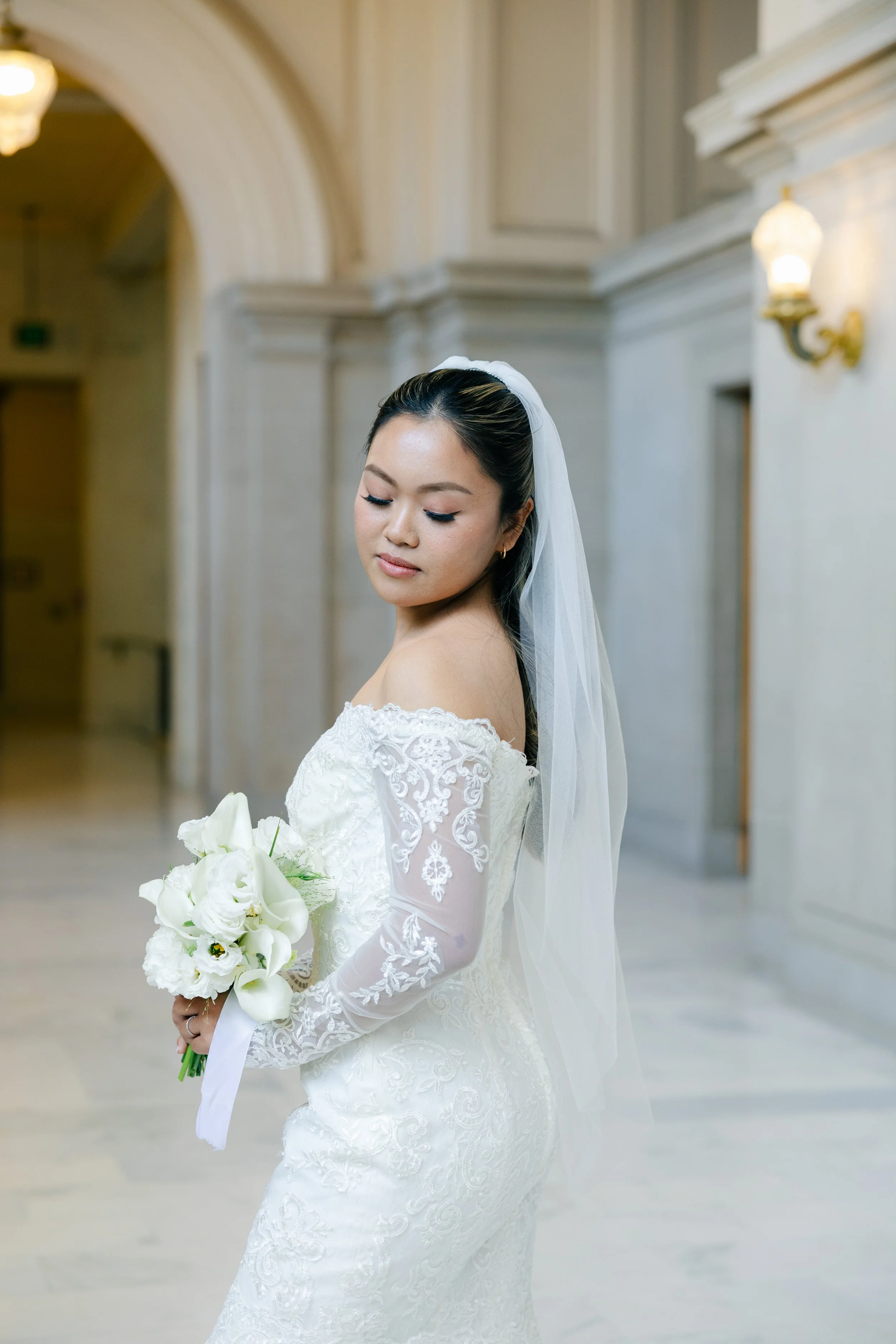 A bride in a white wedding dress holding a bouquet of white flowers, standing in a bright, elegant hall with high ceilings and wall sconces.