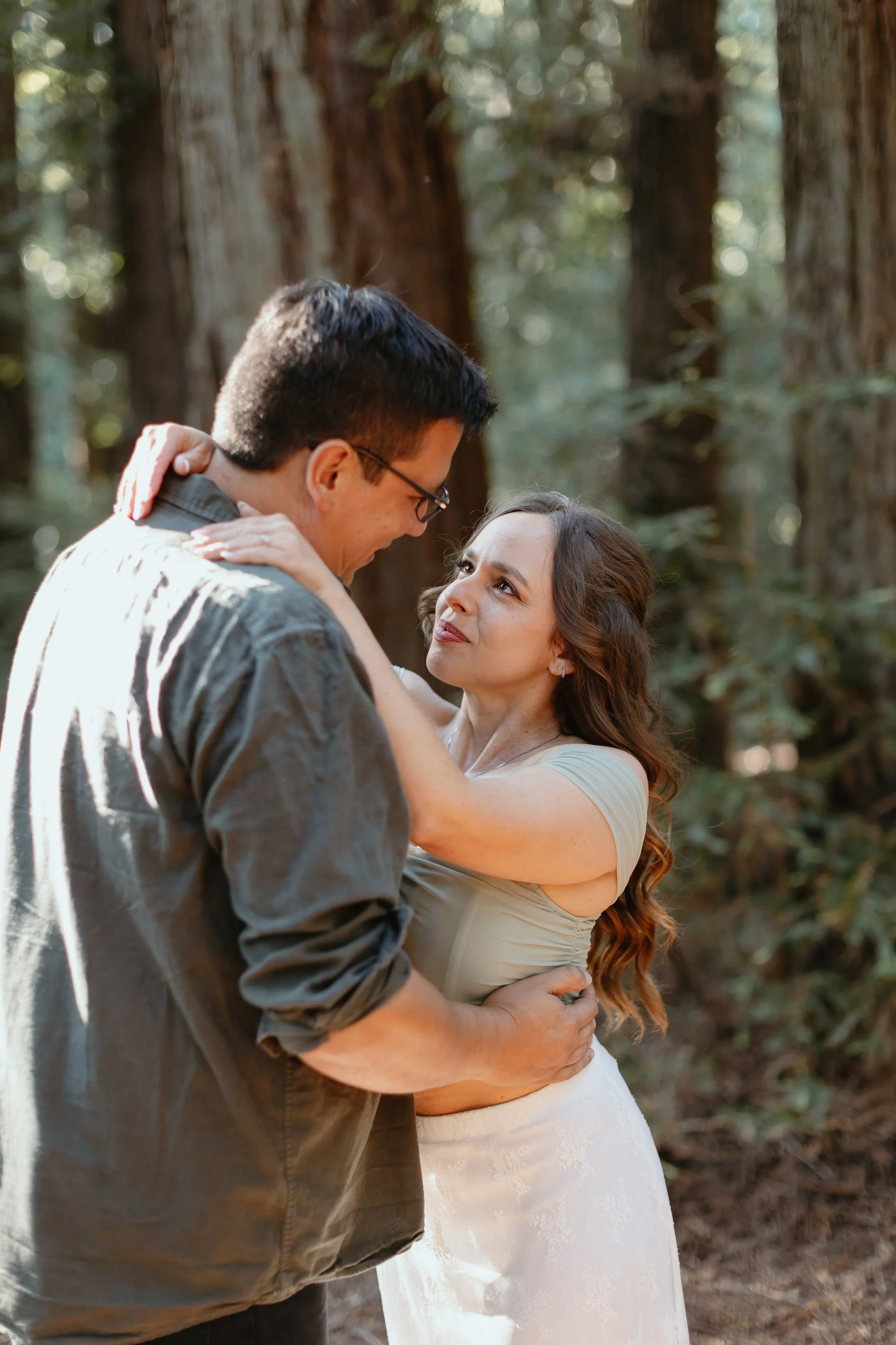 A couple sharing a romantic moment in a forest. The man is wearing glasses and a gray shirt, and the woman is wearing a white dress. They are close, looking into each other's eyes, with the woman's hands on the man's neck and his hands on her waist.