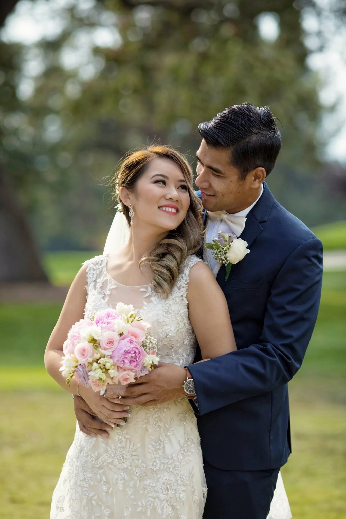 A bride and groom in wedding attire embracing outdoors with trees in the background.