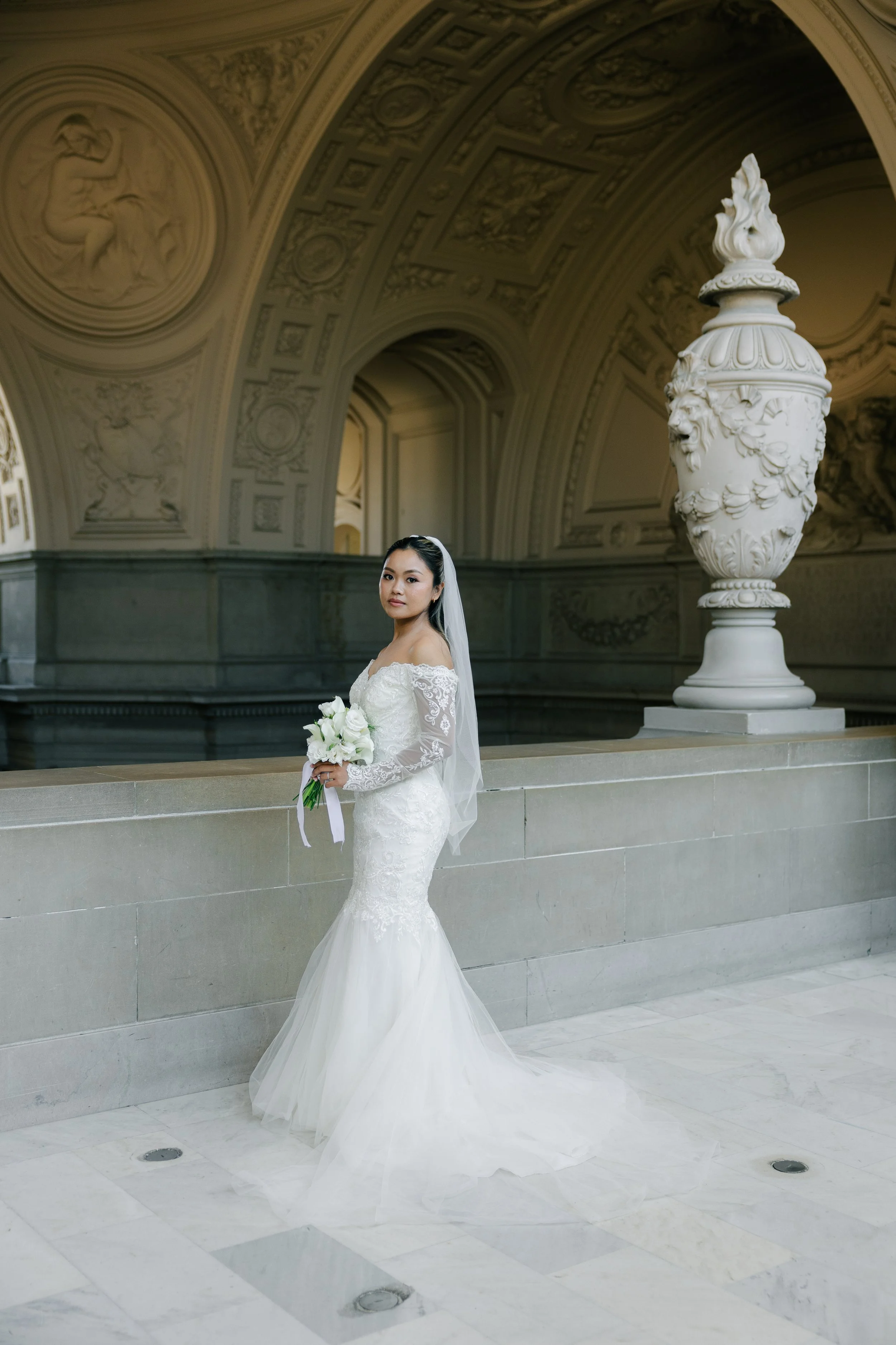 A woman in a white wedding dress holding a bouquet of white flowers standing in an ornate hall with high vaulted ceilings, decorative arches, and classical sculptures.