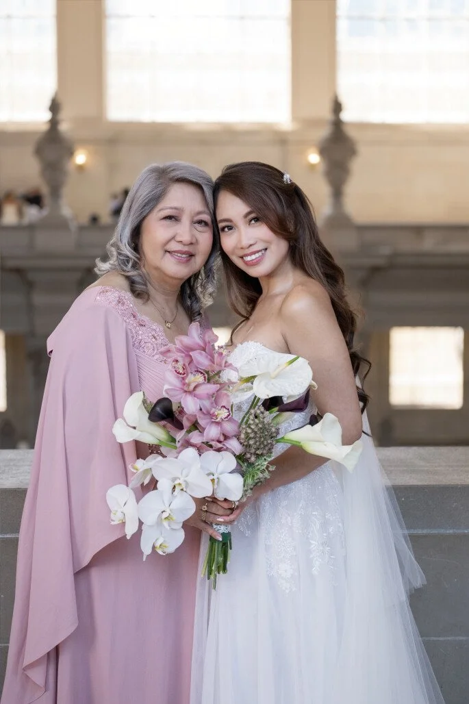 A bride in a white wedding dress holding a bouquet of pink and white flowers, standing next to an older woman in a pink dress, both smiling indoors with large windows in the background.