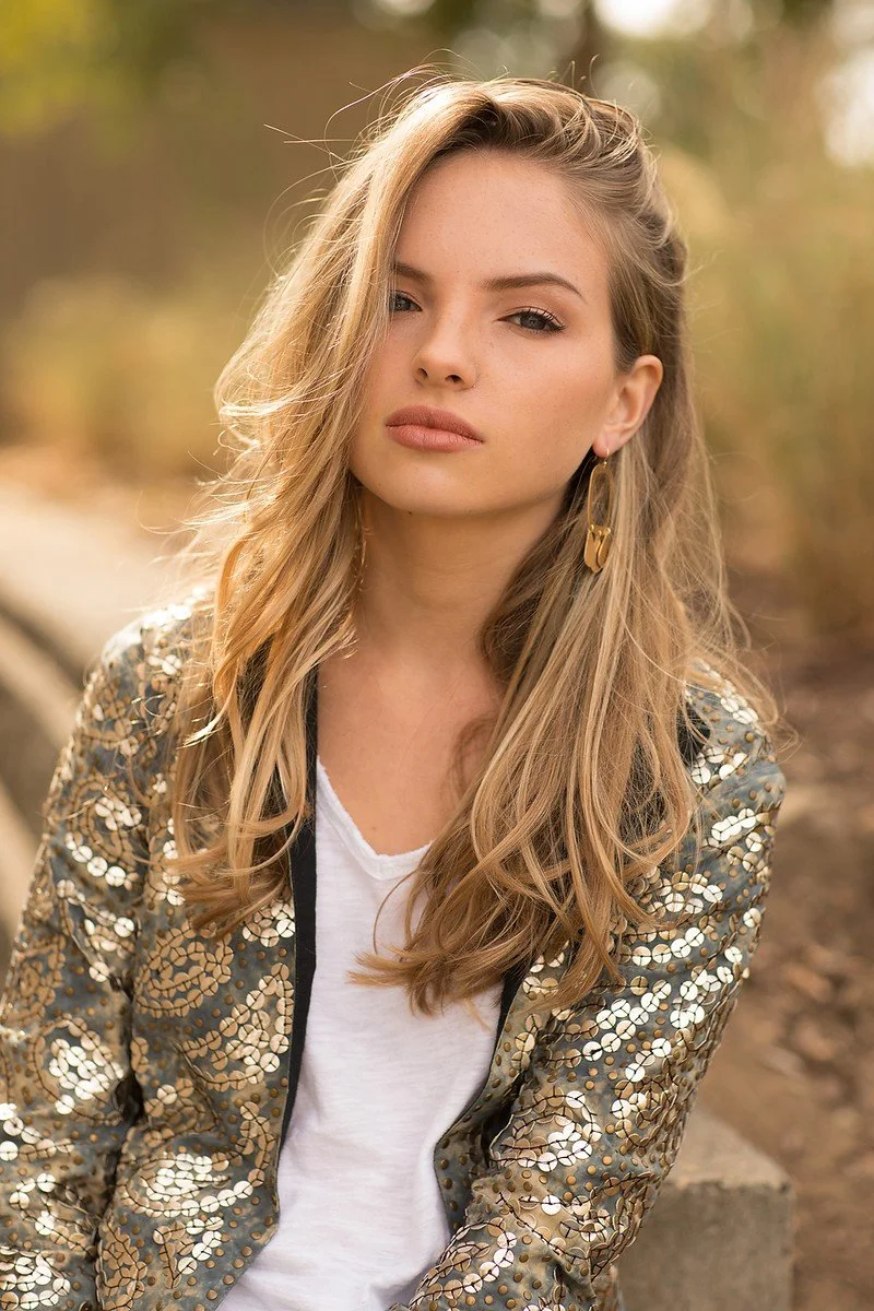 A young woman with long blonde hair and gold earrings sitting outdoors with a blurred background of trees and foliage.