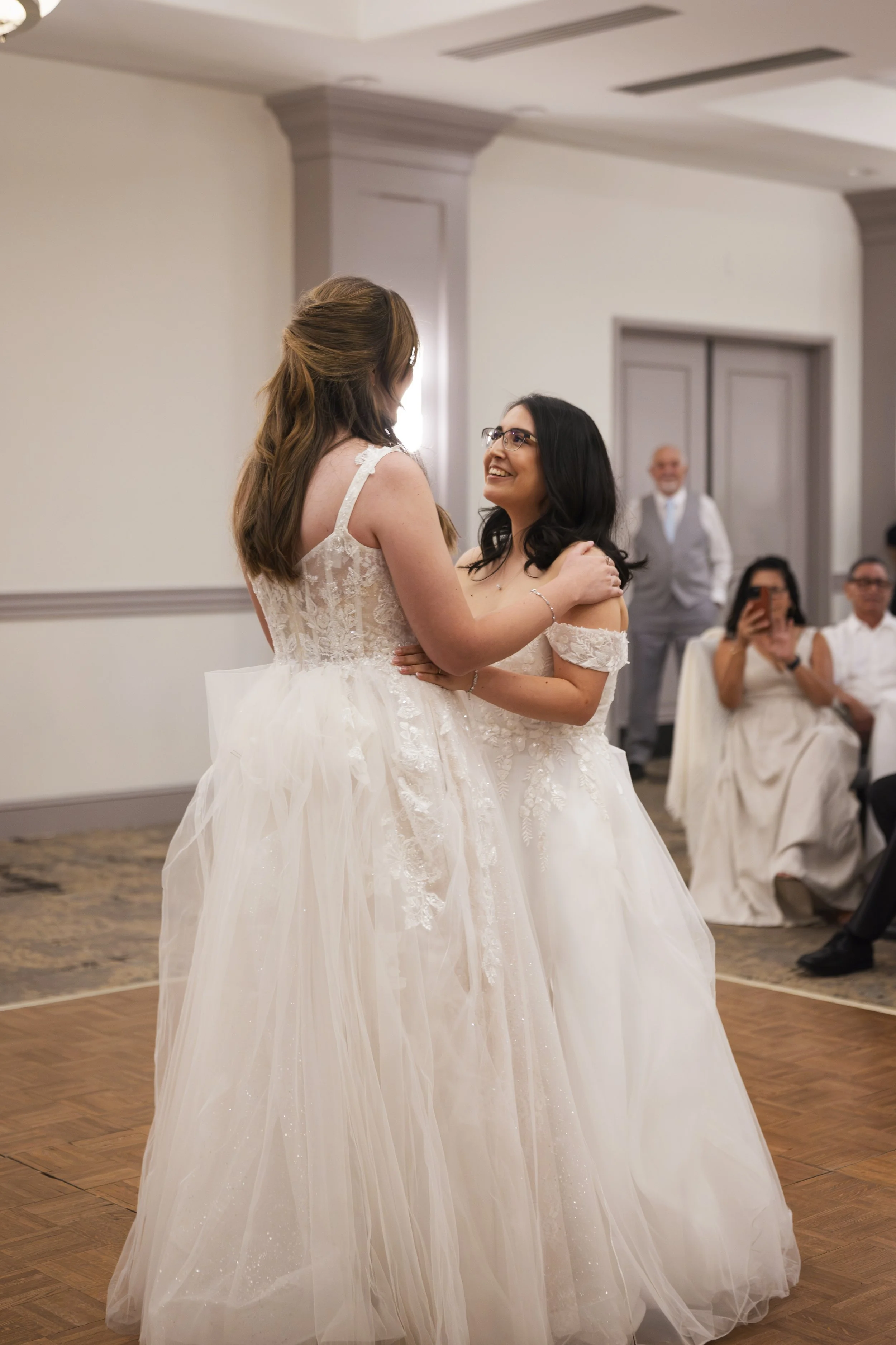Two women in wedding dresses are dancing together at a wedding reception, smiling and holding each other in a ballroom with a wooden dance floor.