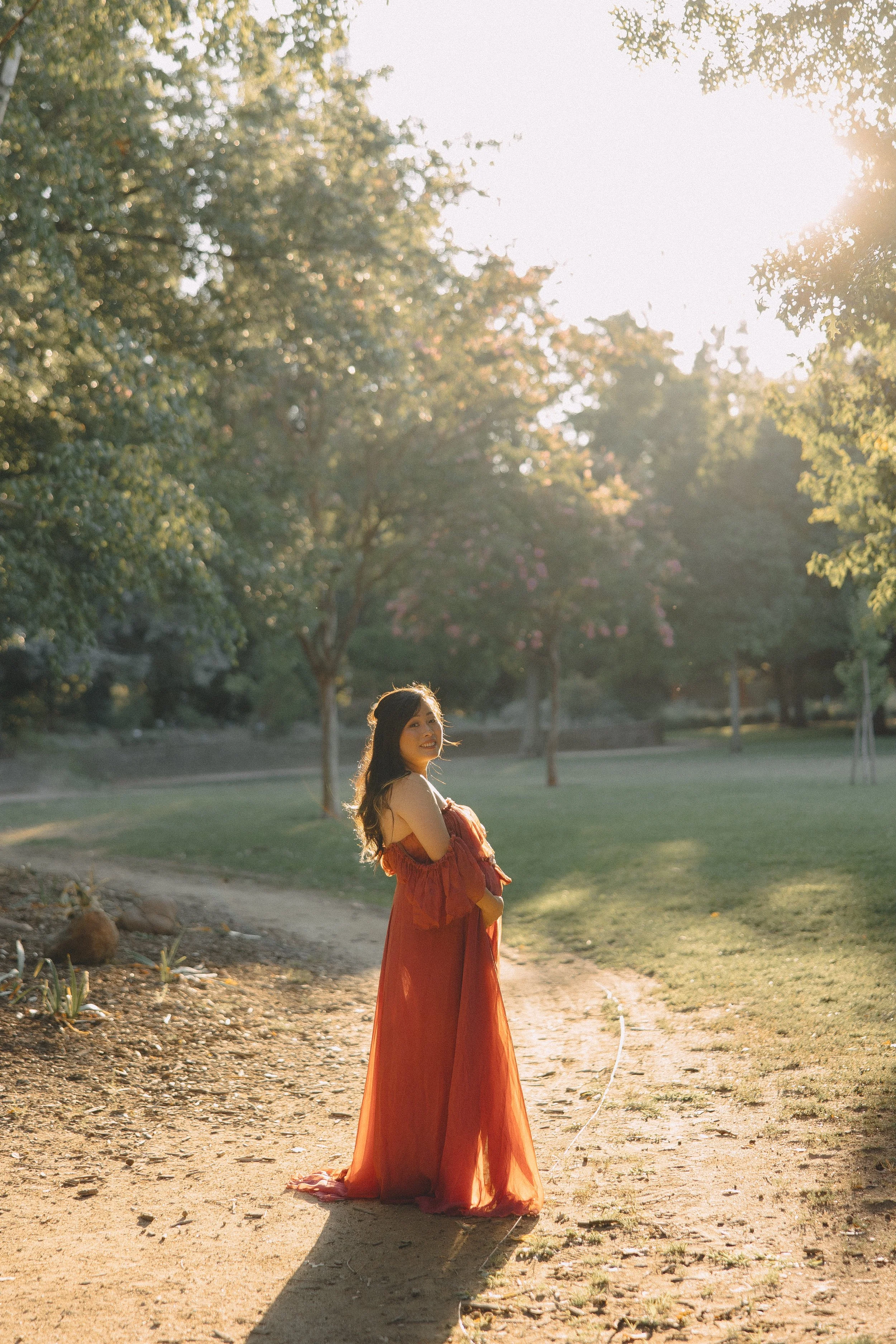A woman in an off-the-shoulder, orange summer dress standing on a dirt path in a park with green grass and trees in the background, illuminated by sunlight.