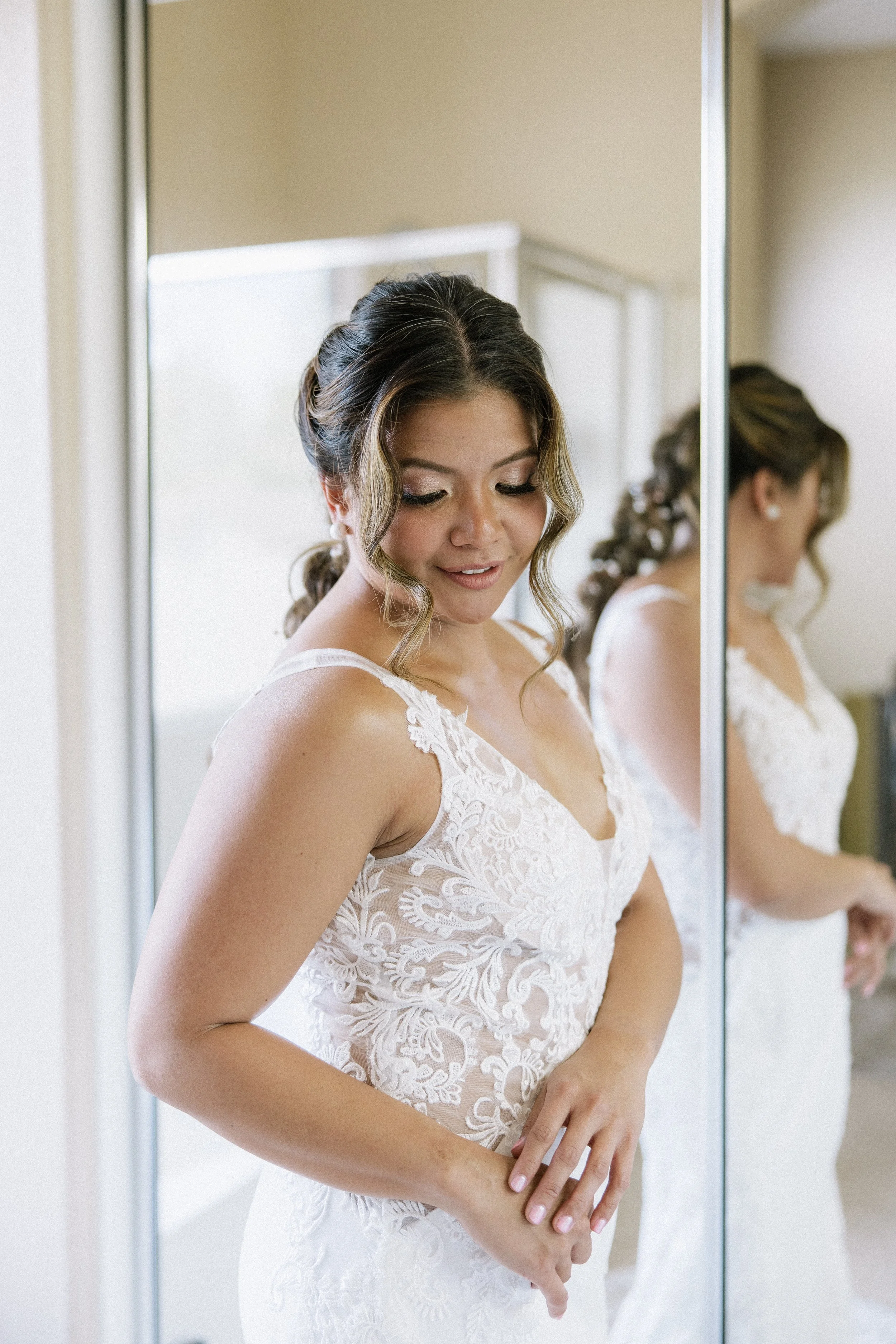A bride standing in front of a mirrored closet door, wearing a white lace wedding dress, with her hair styled in loose curls and looking down with a gentle smile.