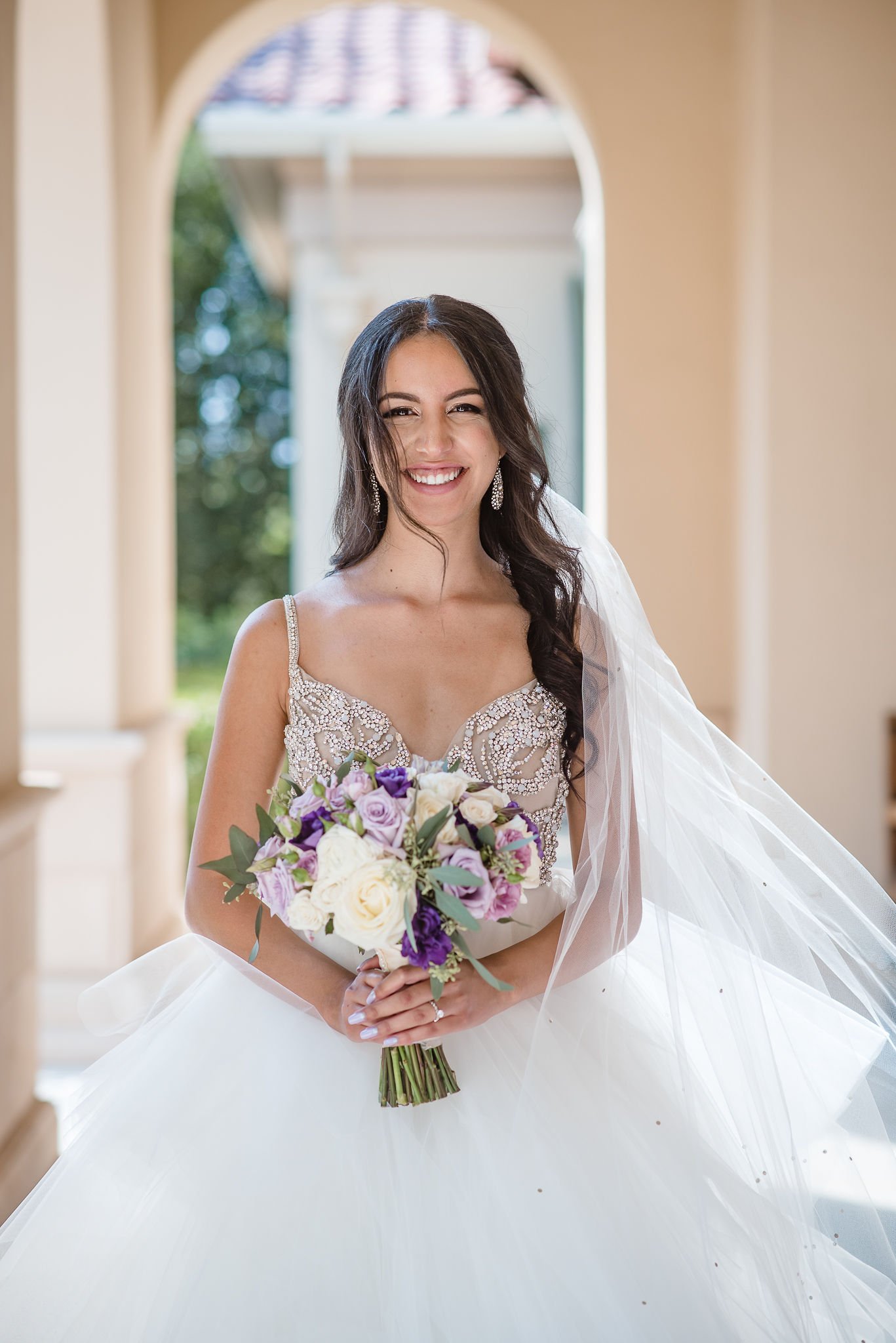 A smiling bride in a wedding gown holding a bouquet of purple, white, and green flowers indoors with an arched window in the background.