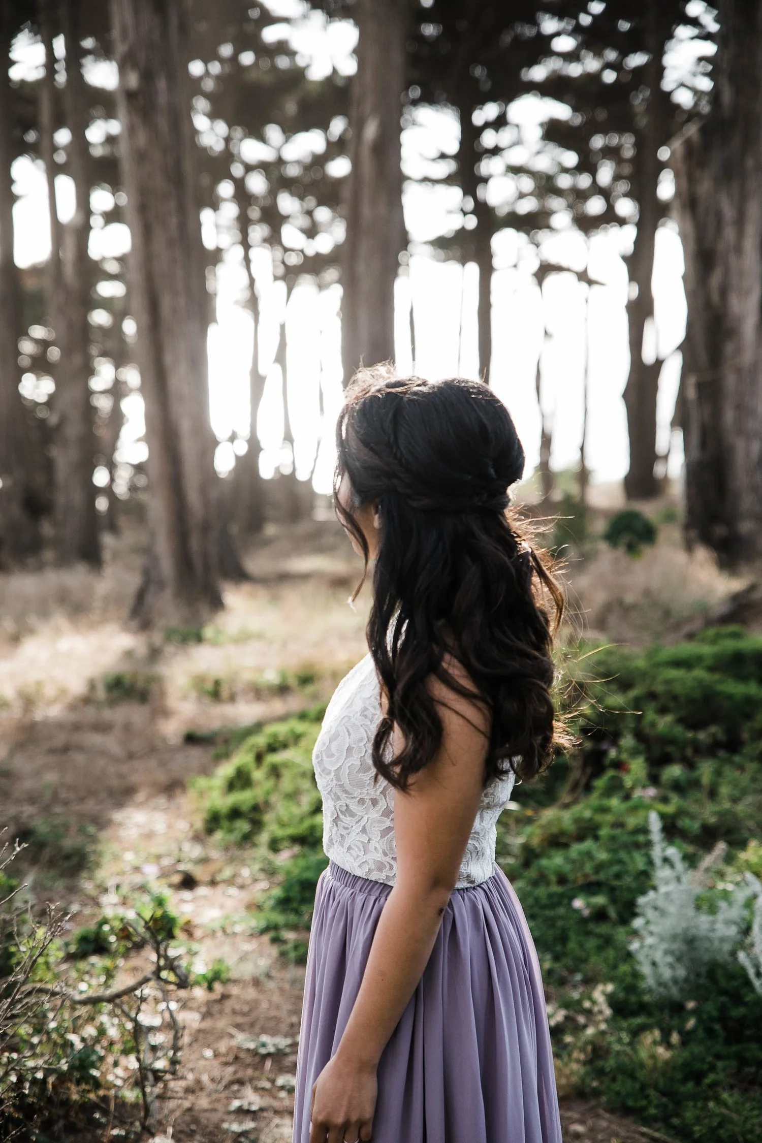 A woman in a white lace top and a long purple skirt standing in a wooded area with tall trees, facing sideways with her dark, wavy hair flowing down her back, backlit by sunlight.