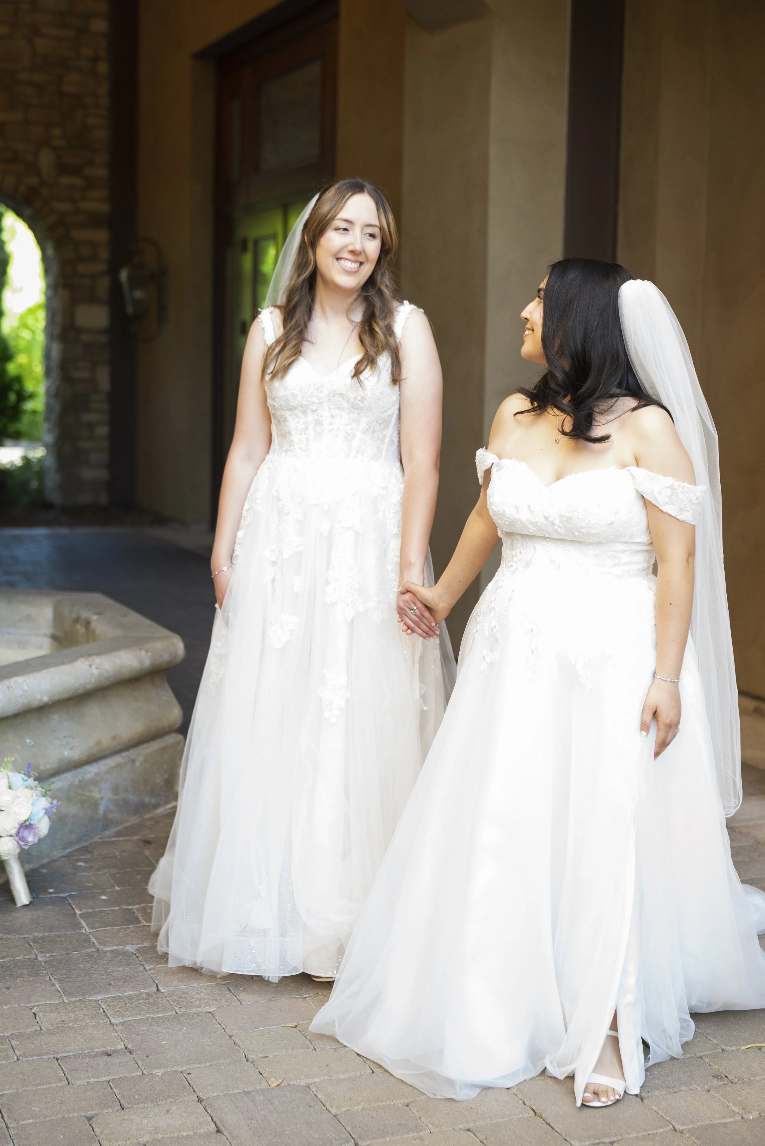 Two brides standing outdoors, holding hands and smiling at each other, dressed in white wedding gowns and veils.