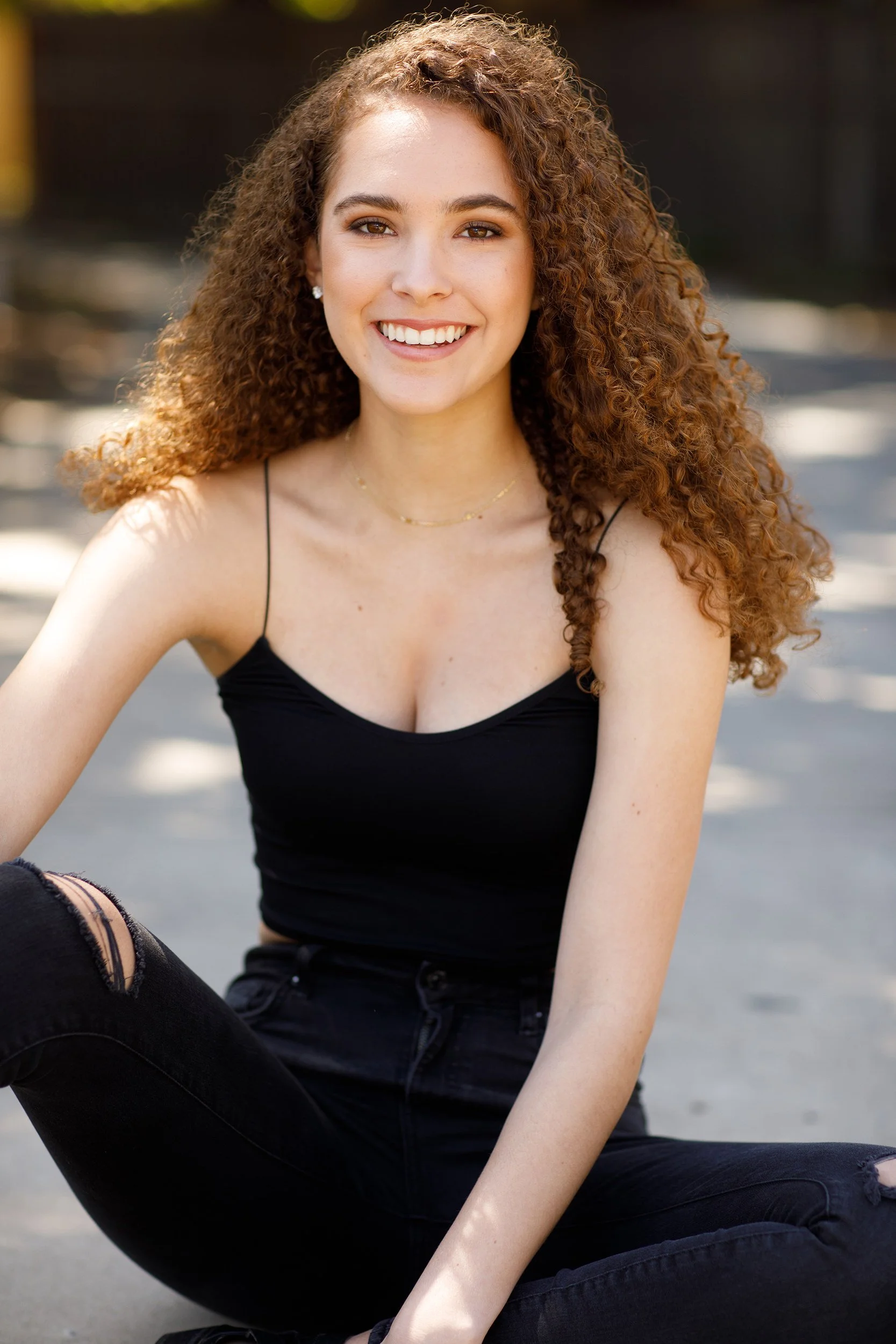 A young woman with curly red hair smiling outdoors, wearing a black spaghetti strap top and ripped black jeans.