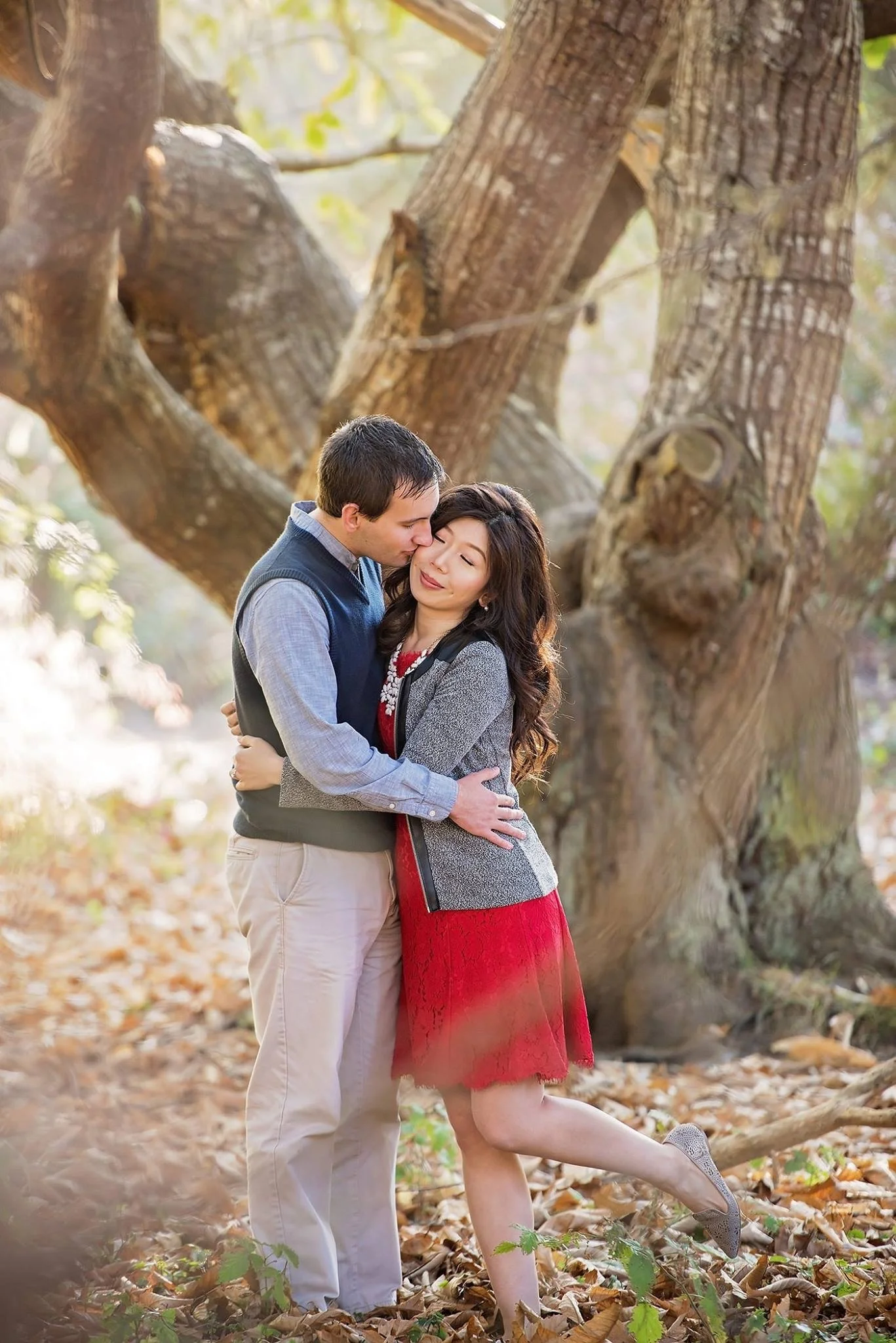 A couple hugging and sharing a kiss in a wooded area with fallen leaves, a large tree with a textured trunk in the background, and sunlight filtering through the trees.