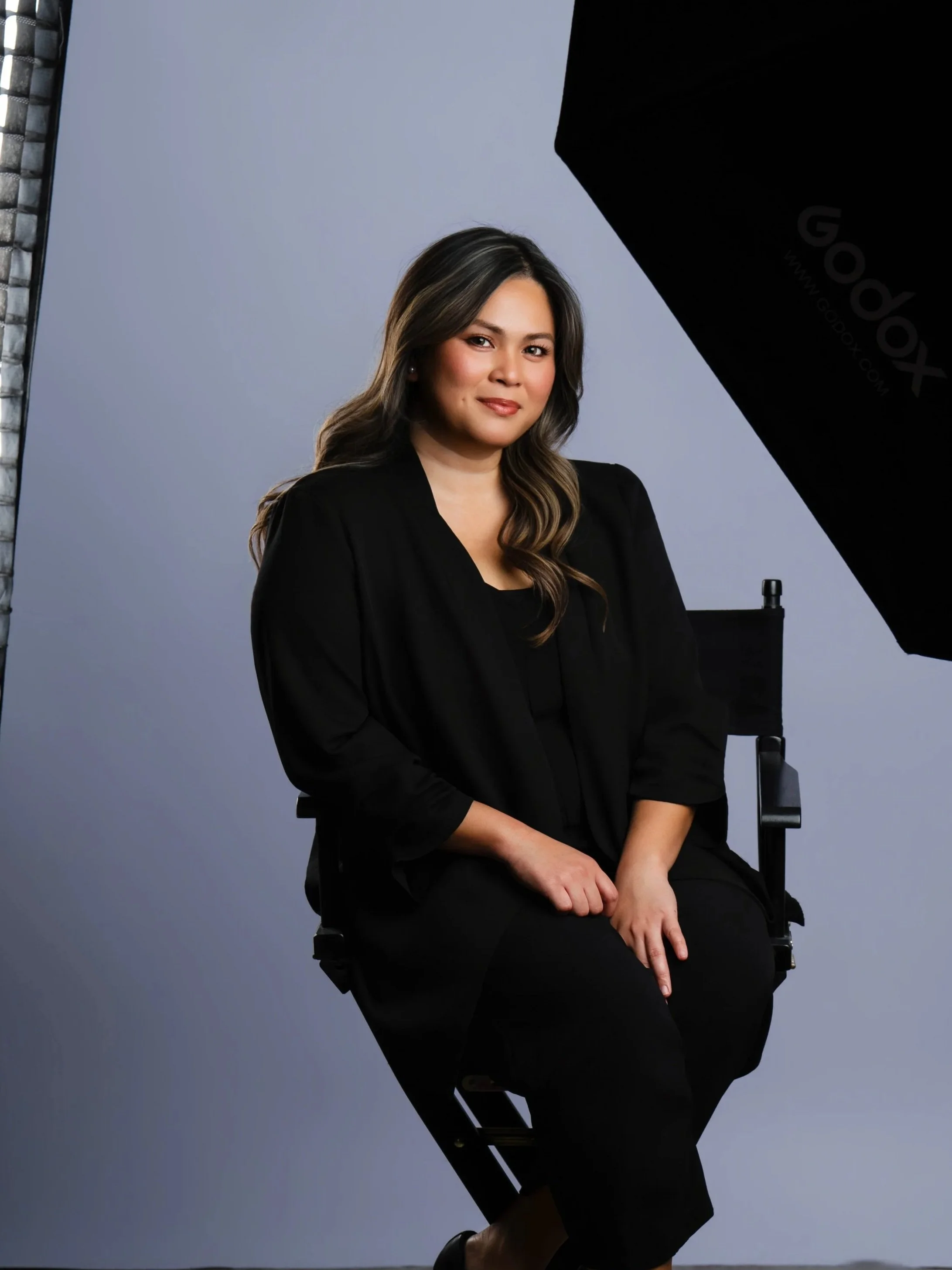 A woman with long wavy hair, dressed in black, sitting on a black director's chair in a photo studio, with lighting equipment and a plain background.