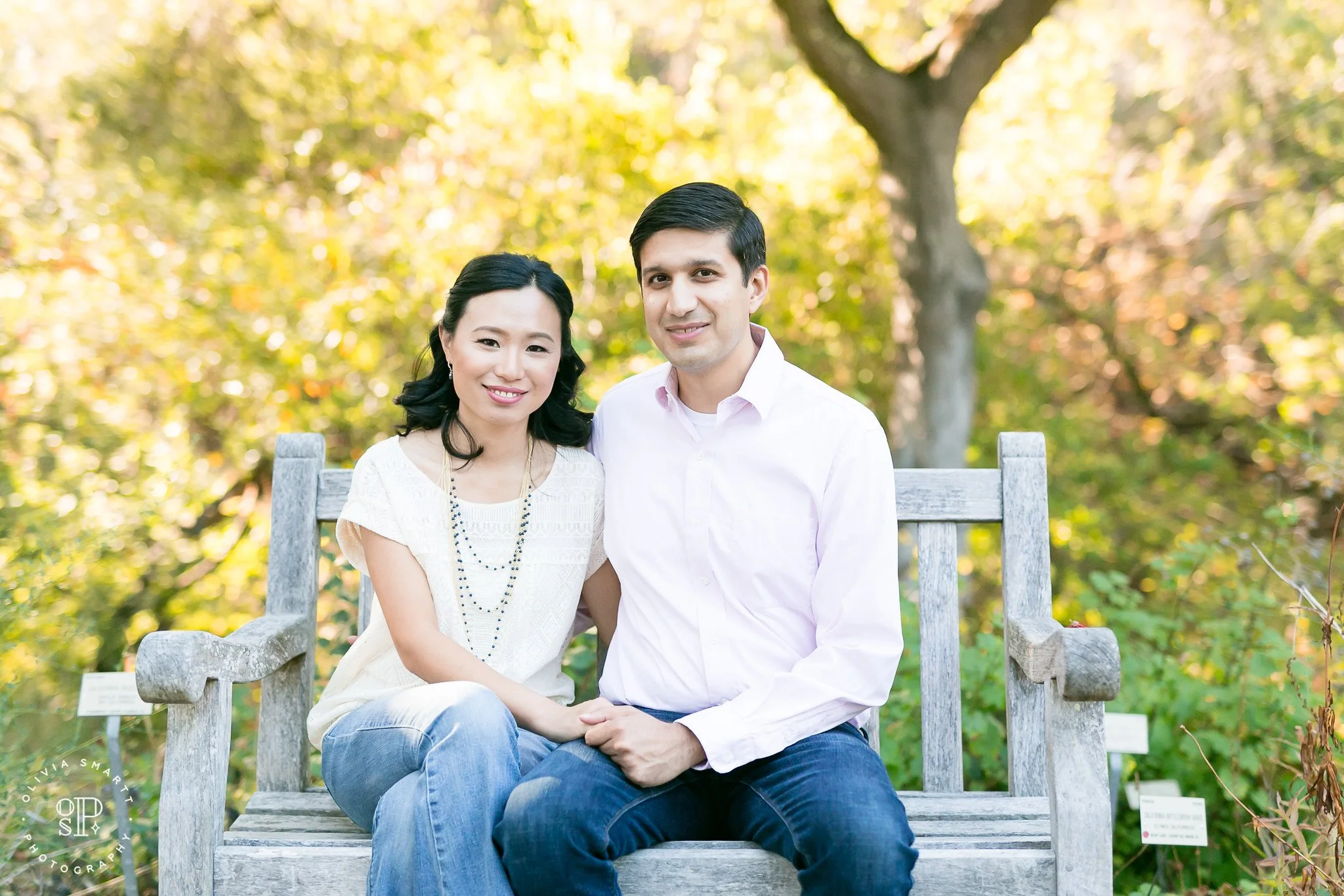 A smiling couple sitting on a wooden bench outdoors surrounded by green and yellow foliage during daytime.