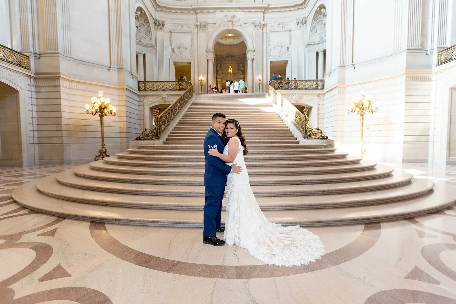A newlywed couple in wedding attire, standing on a grand staircase inside a luxurious building with ornate decor. The bride is wearing a white lace wedding gown with a train, and the groom is in a blue suit. The staircase has golden railings, and the