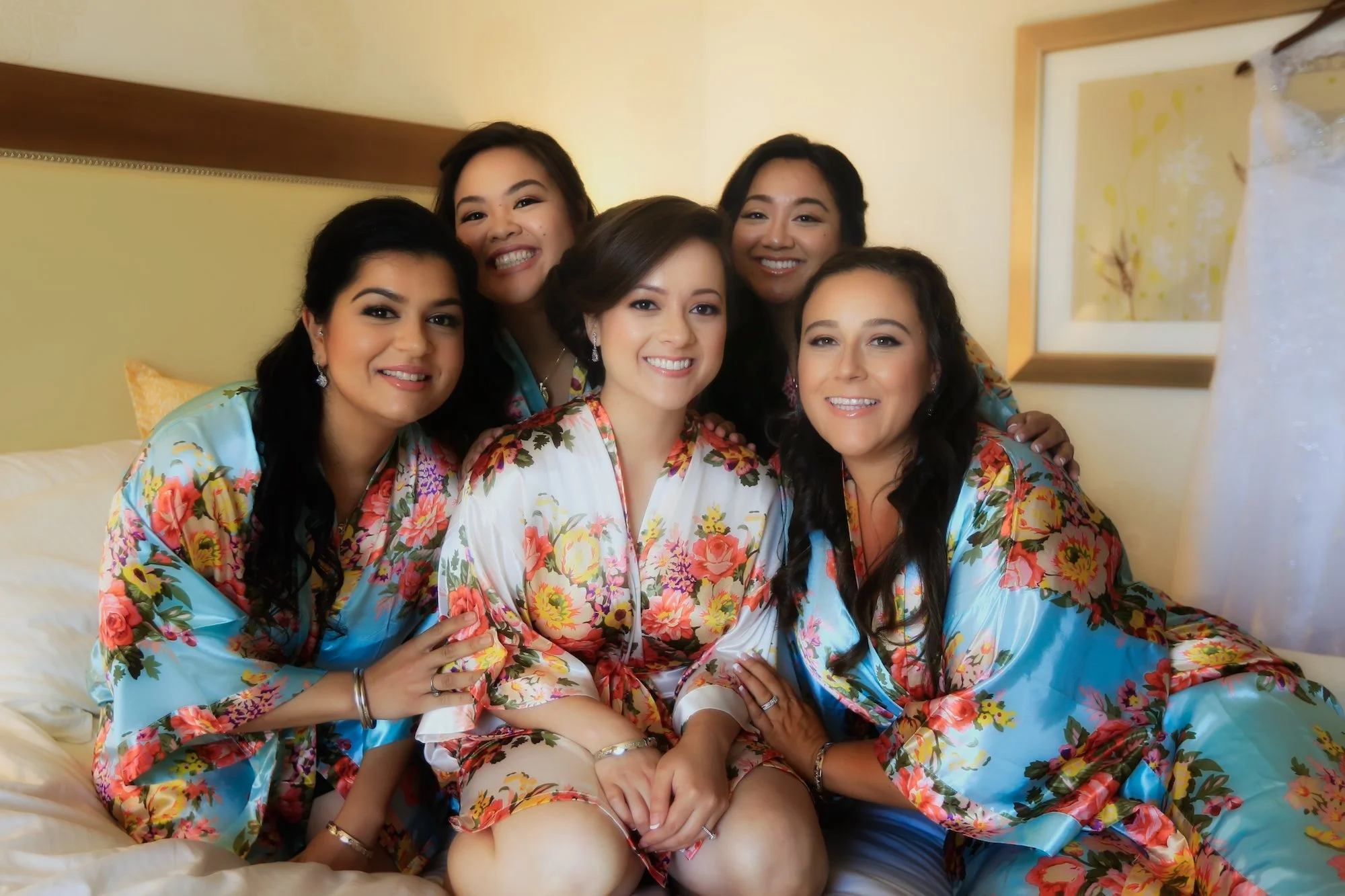 Group of six women wearing matching floral robes, smiling and sitting together on a bed in a hotel room.