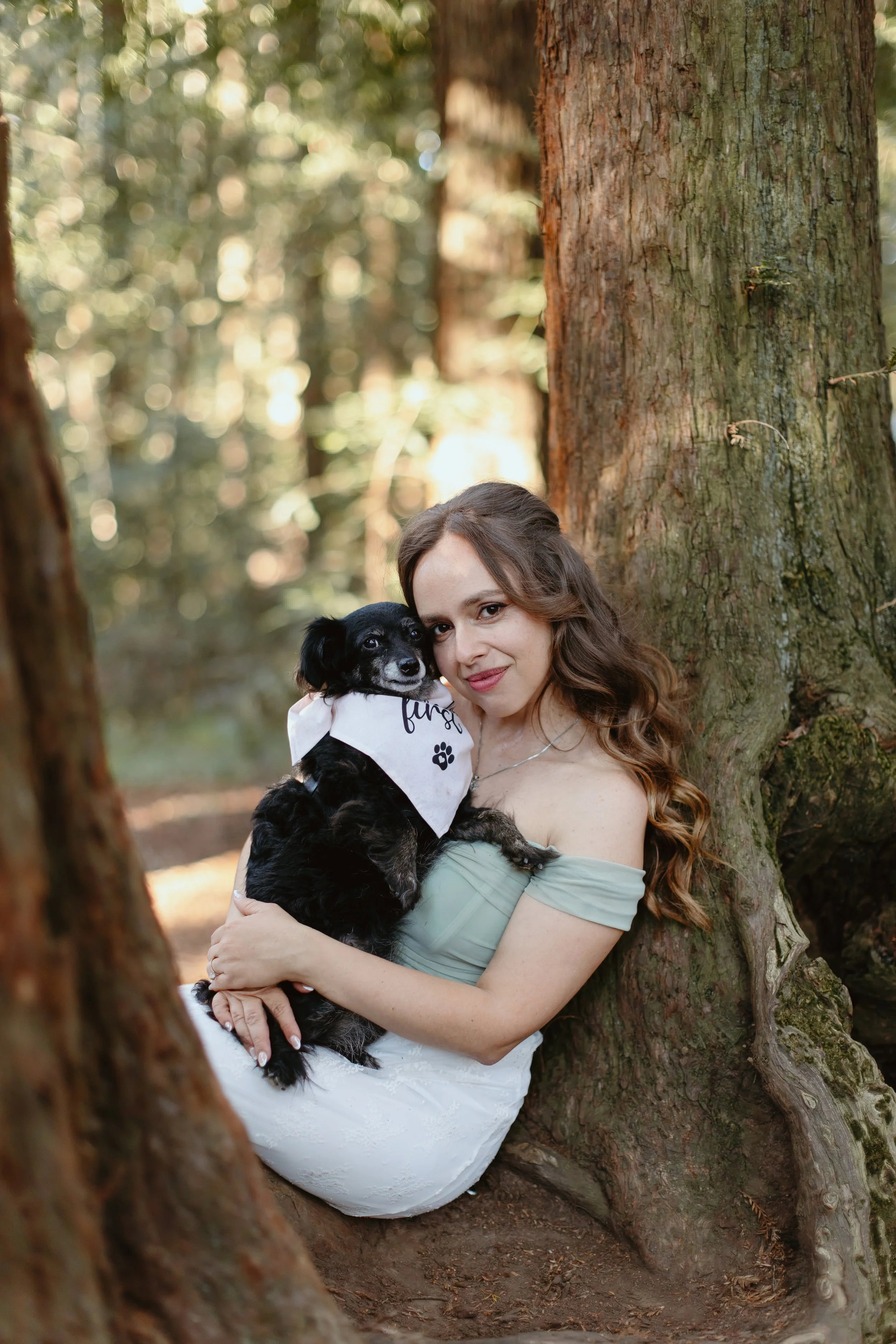 A woman with brown hair sitting in a hollowed out tree trunk in a forest, holding a black and white dog wearing a bandana.