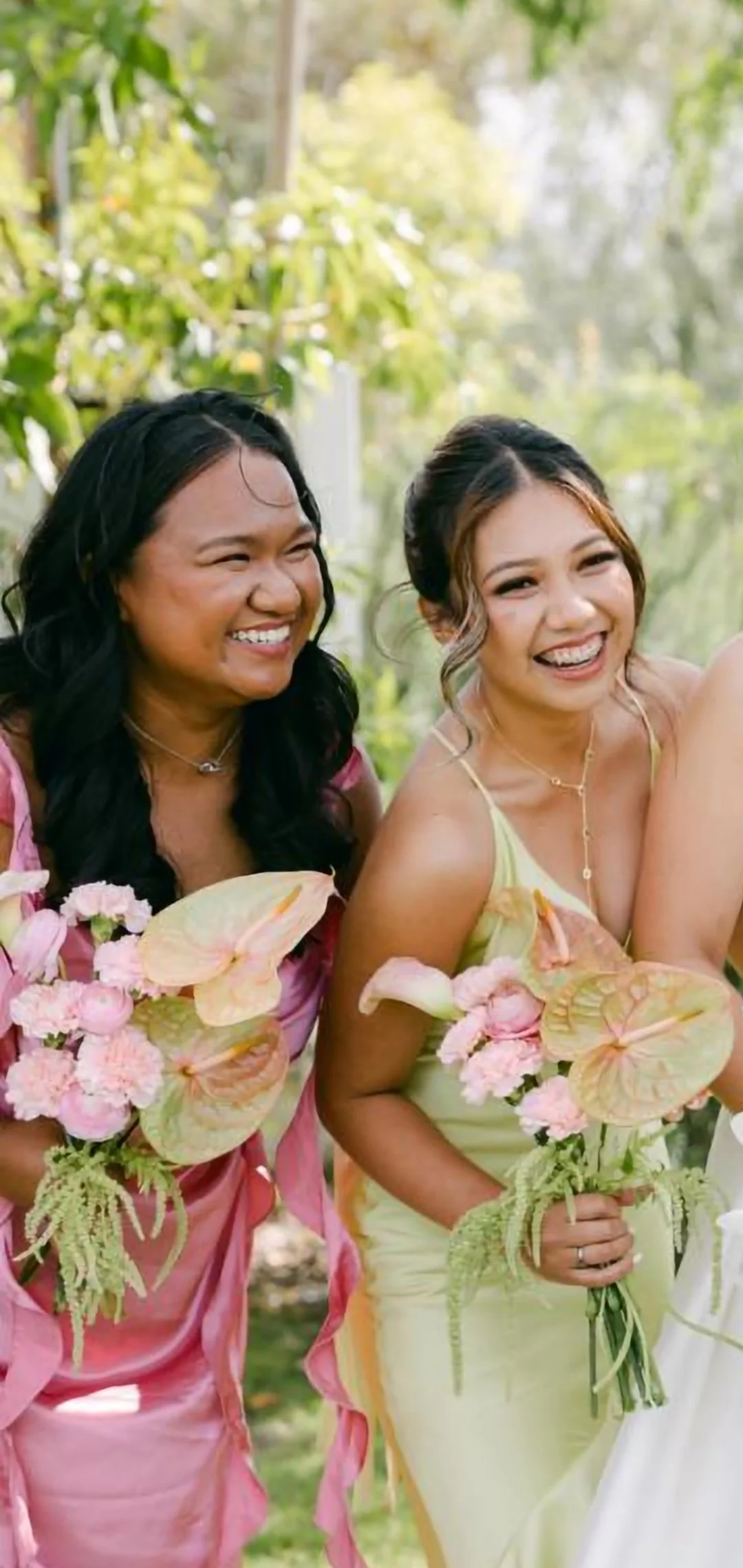 Two women smiling and holding bouquets of pink flowers outdoors, surrounded by greenery.