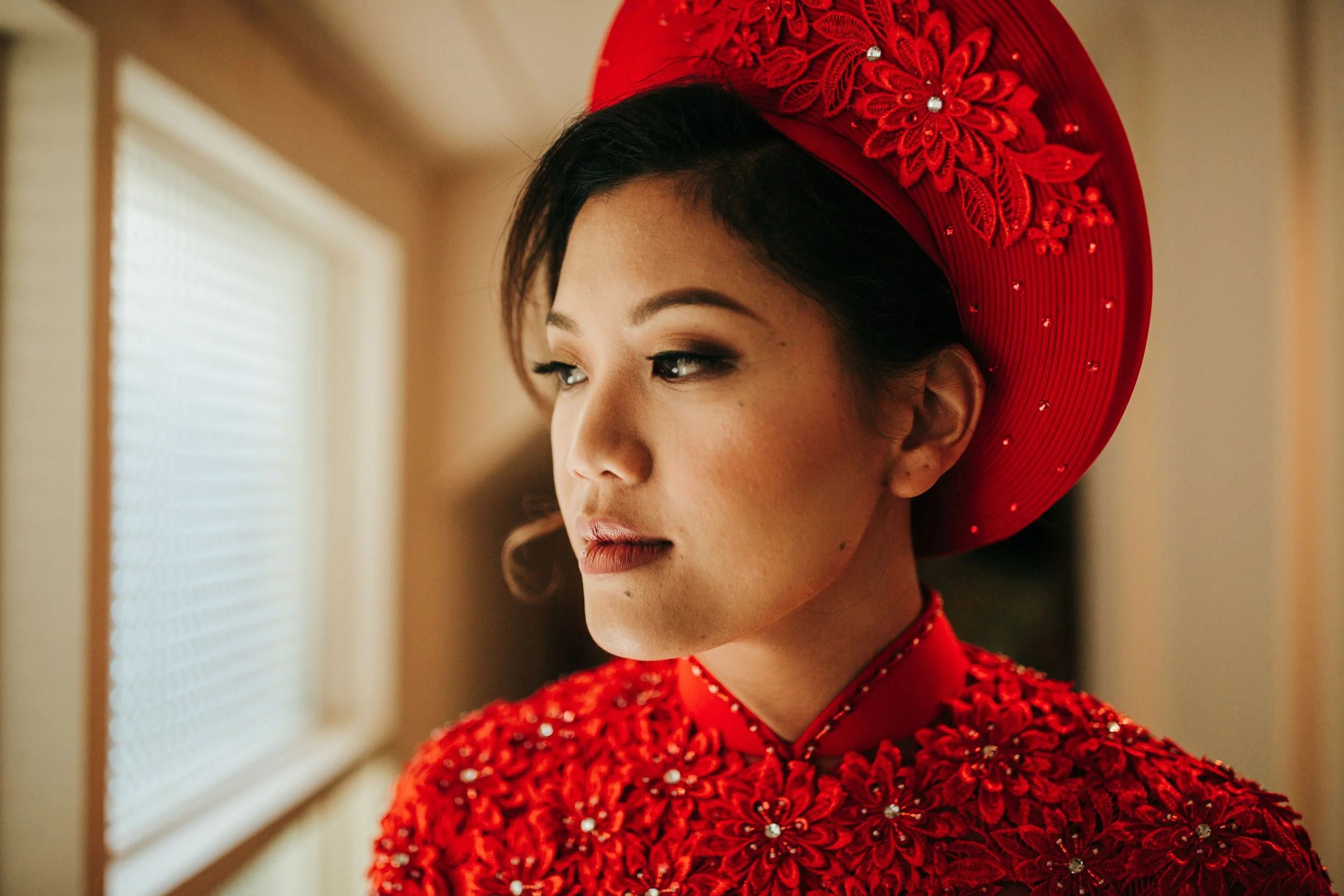 A woman dressed in a red embroidered traditional outfit and matching wide-brimmed hat, looking thoughtfully to the side near a window.