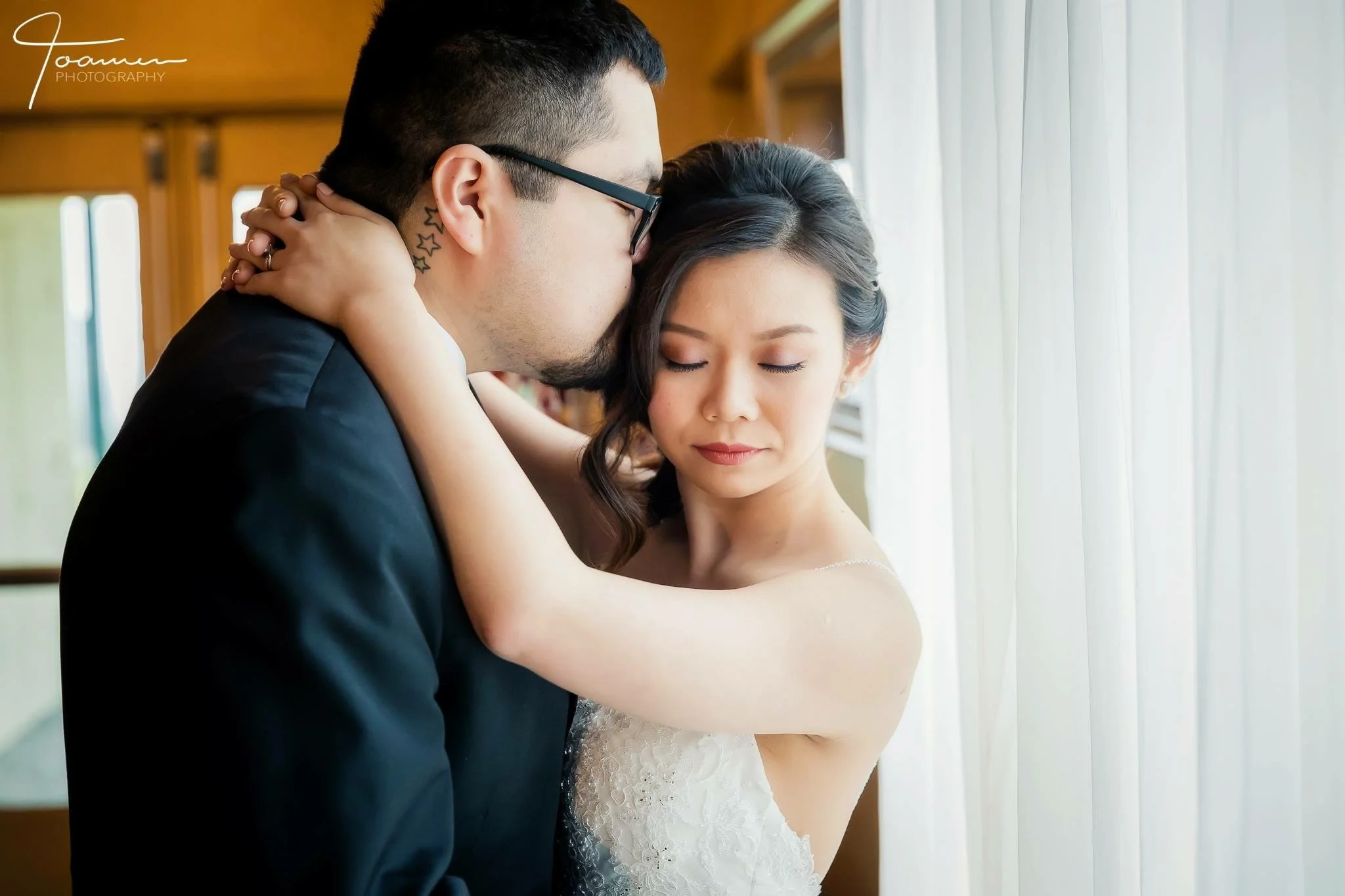 A bride and groom sharing a tender moment indoors, with natural light coming through curtains.