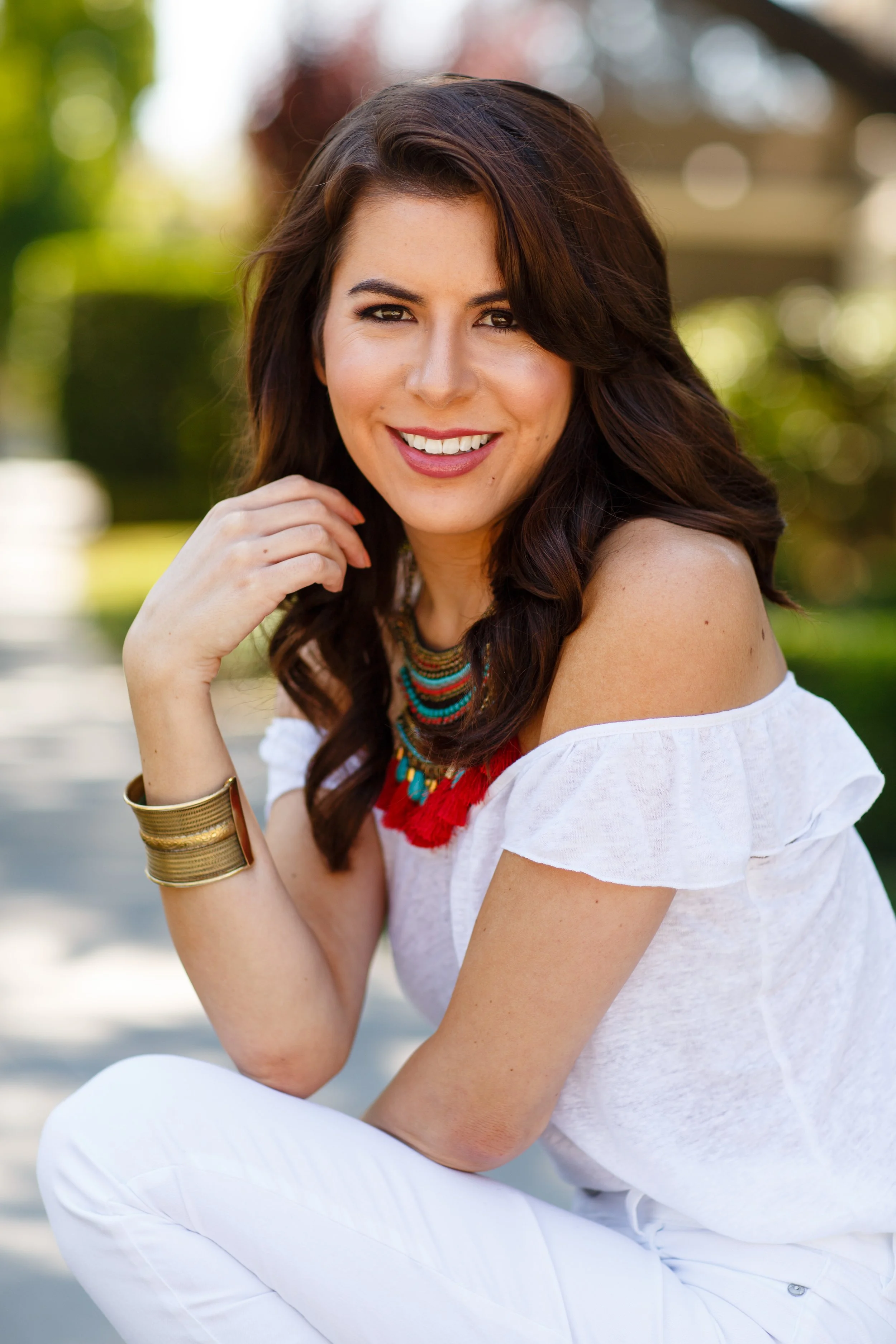 A woman with long brown hair, wearing a white off-shoulder top, colorful necklace, and gold bracelet, sitting outdoors and smiling.