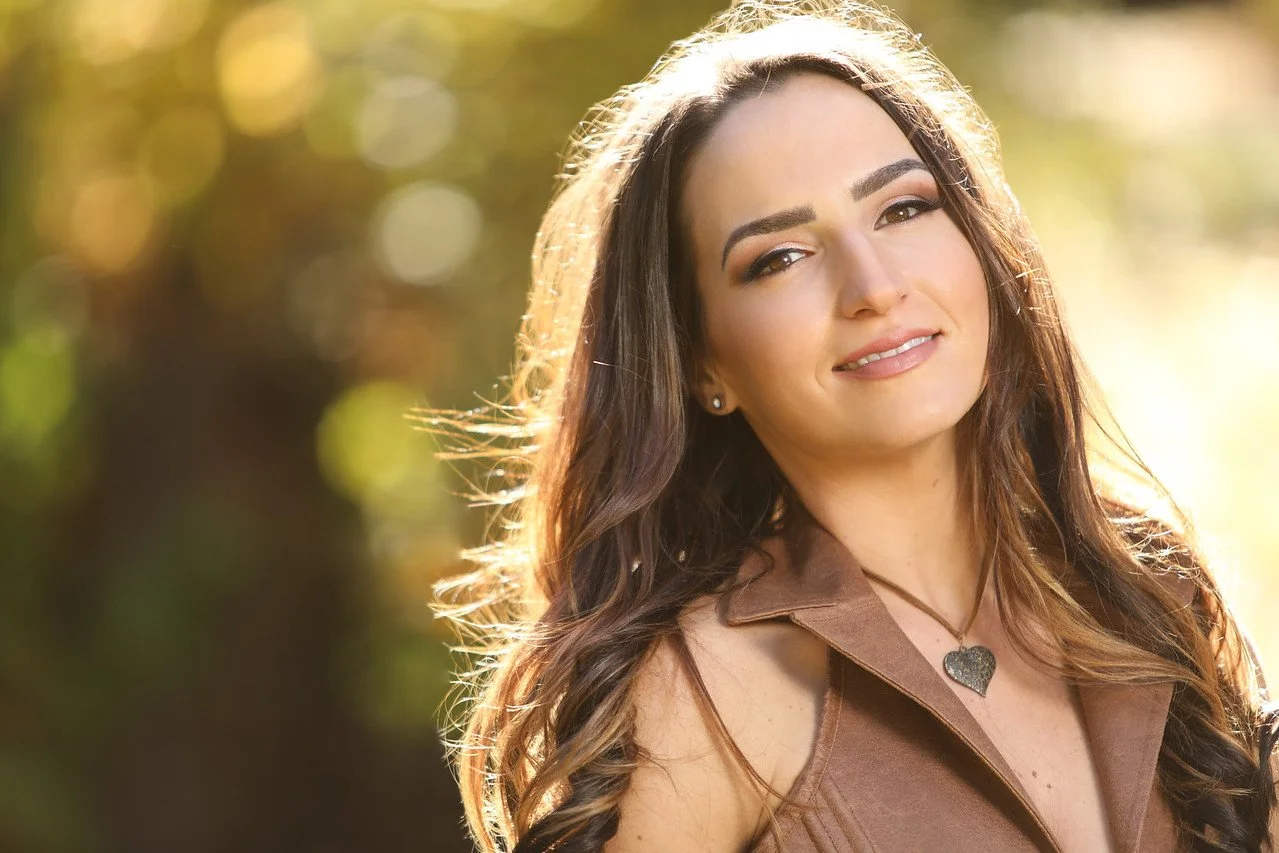 Young woman with long, wavy brown hair smiling outdoors in warm sunlight, wearing a brown sleeveless top and a heart necklace.