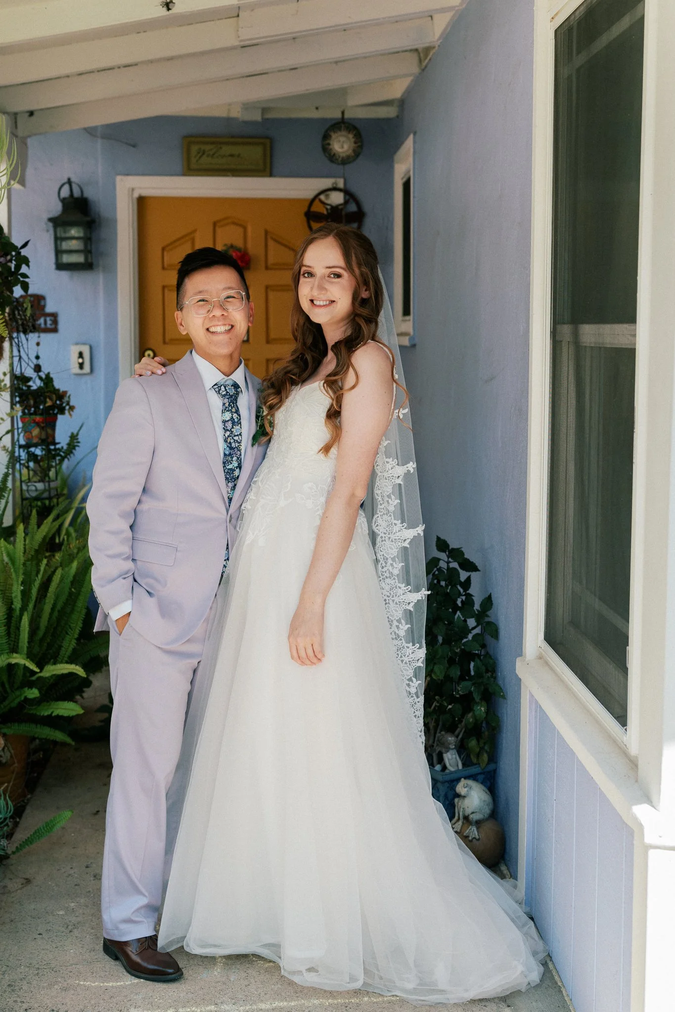 A smiling bride and groom stand together outside a house, the groom wearing a light purple suit and the bride in a white wedding gown. The background includes a yellow front door, a blue house wall, plants, and decorative items.