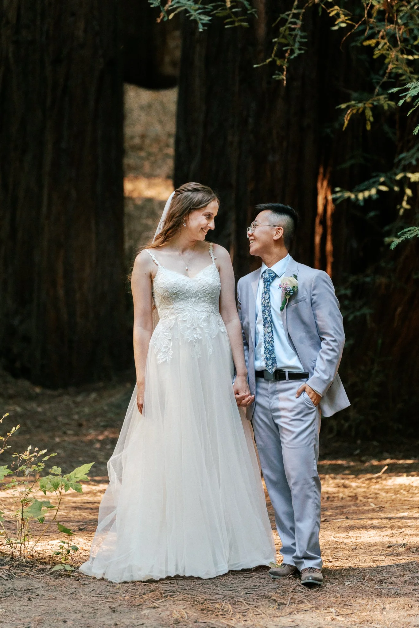 A bride and groom holding hands and smiling at each other in a forest setting, with tall trees and sunlight filtering through the leaves.