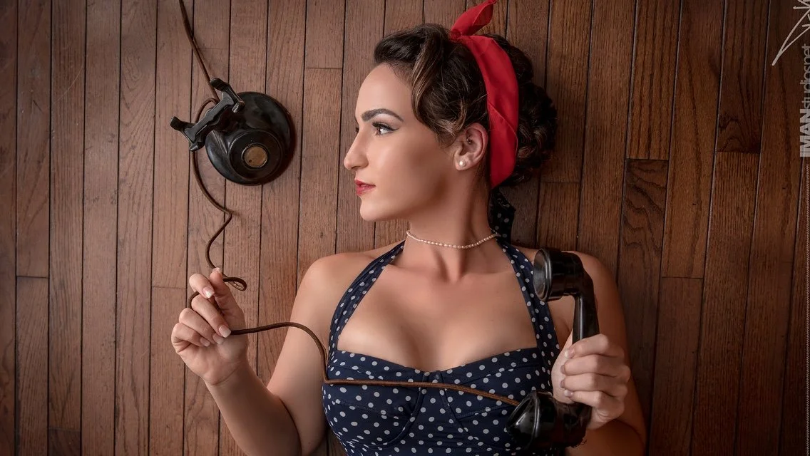 A woman with vintage style hair and makeup, wearing a polka dot dress and red headscarf, lying on a wooden floor holding an old-fashioned rotary phone. She looks to the side with a contemplative expression.