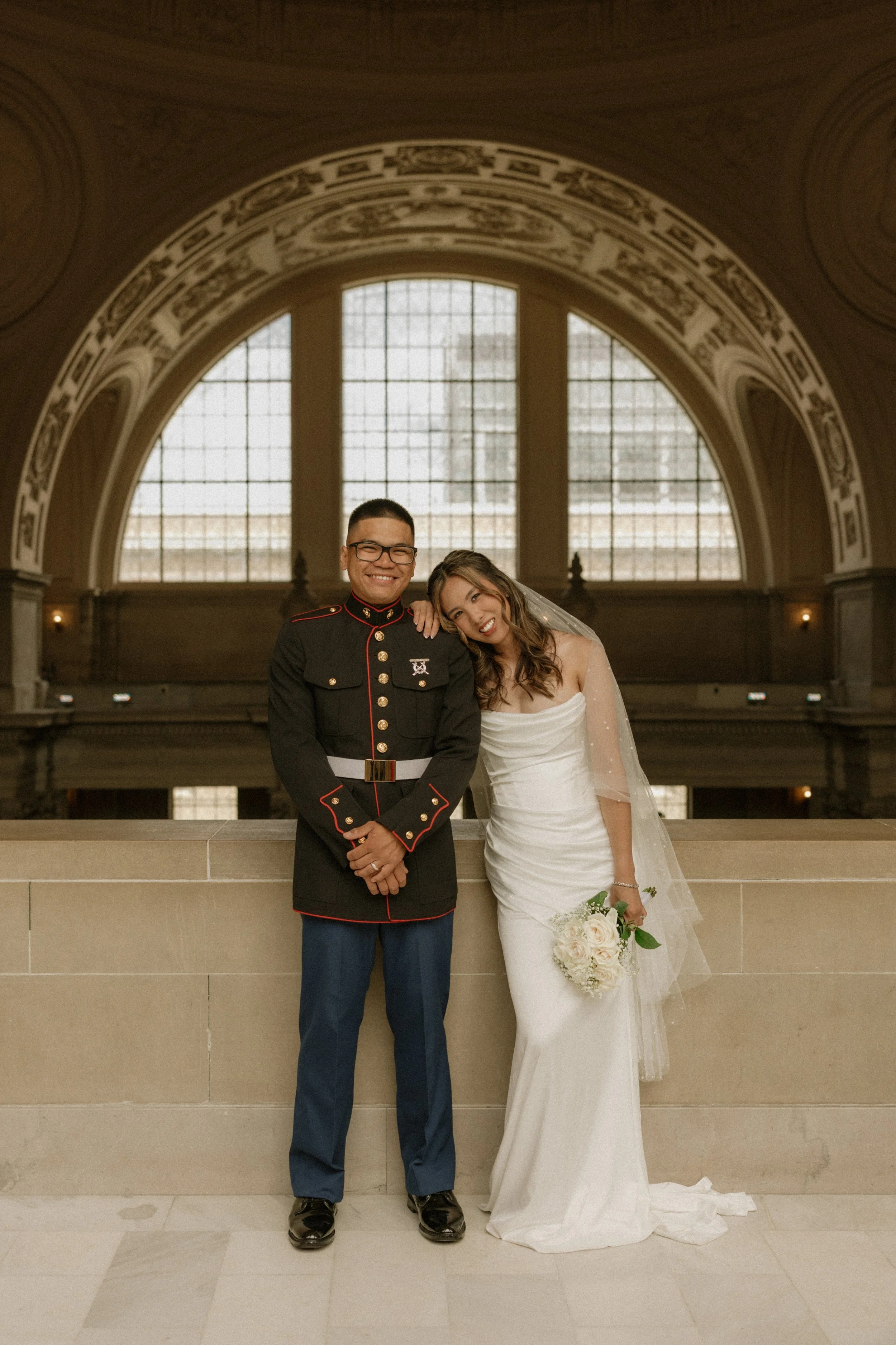 A couple, a man in military uniform and a woman in a white wedding dress, standing together inside a large hall with high arched windows and ornate ceiling details. They are smiling, and the woman is holding a bouquet of white roses.