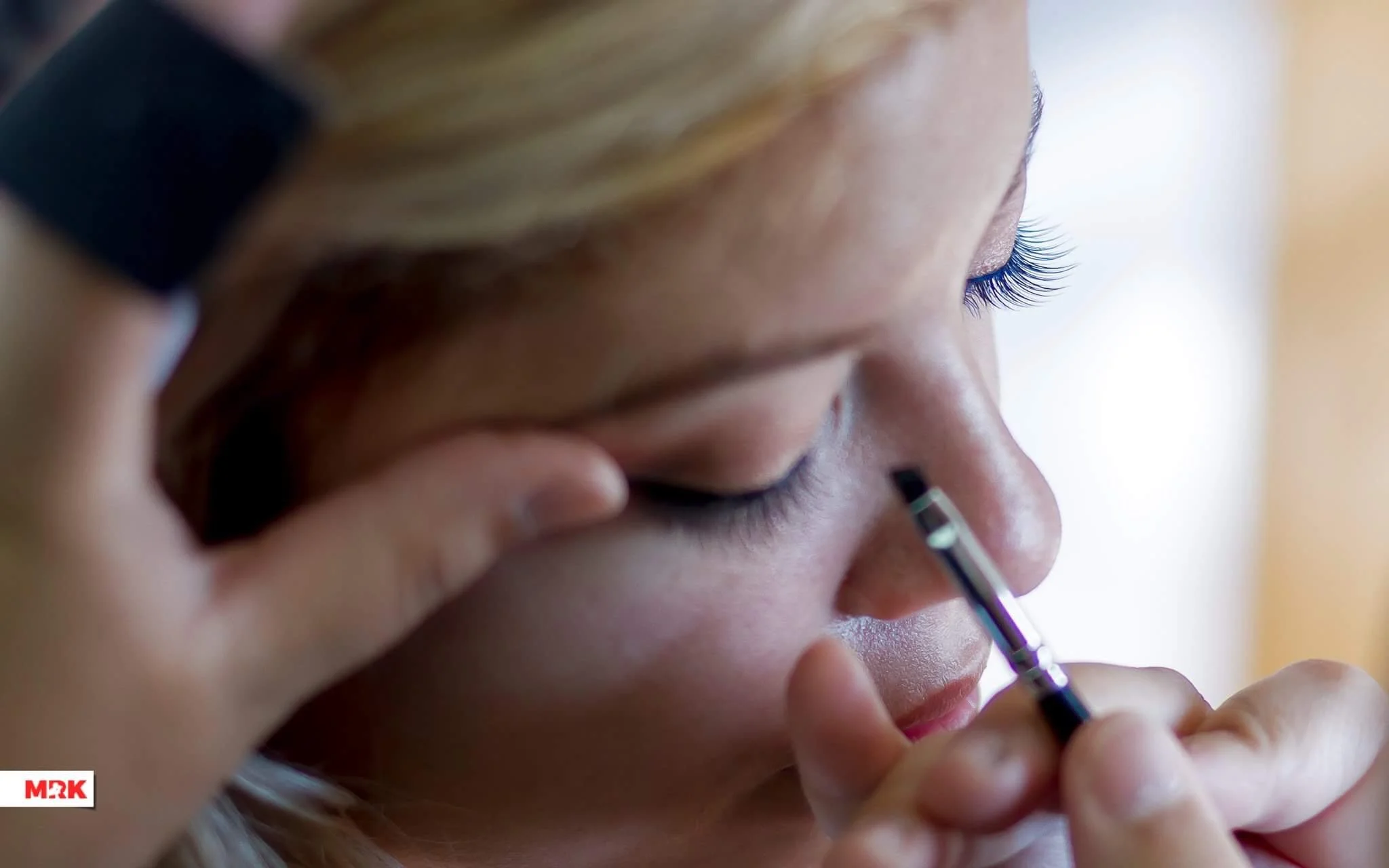 A woman with blonde hair getting her makeup done, applying eyeliner with a brush close to her eye.