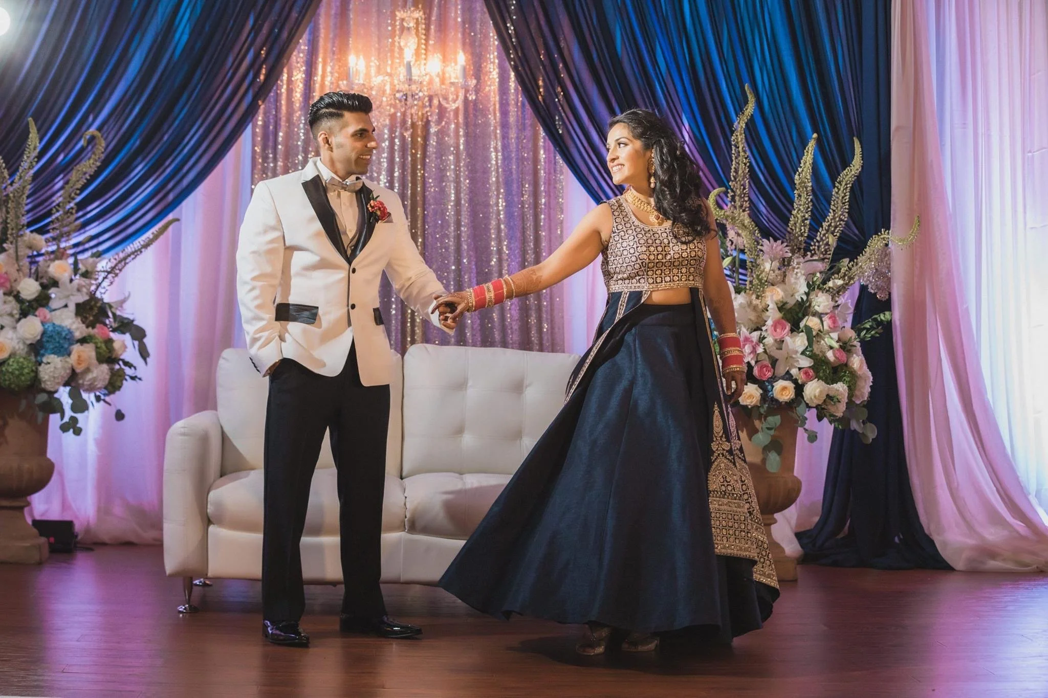 A couple dancing at their wedding ceremony on a decorated stage with flowers, curtains, and a chandelier, with the woman wearing a traditional Indian outfit and the man in a tuxedo.