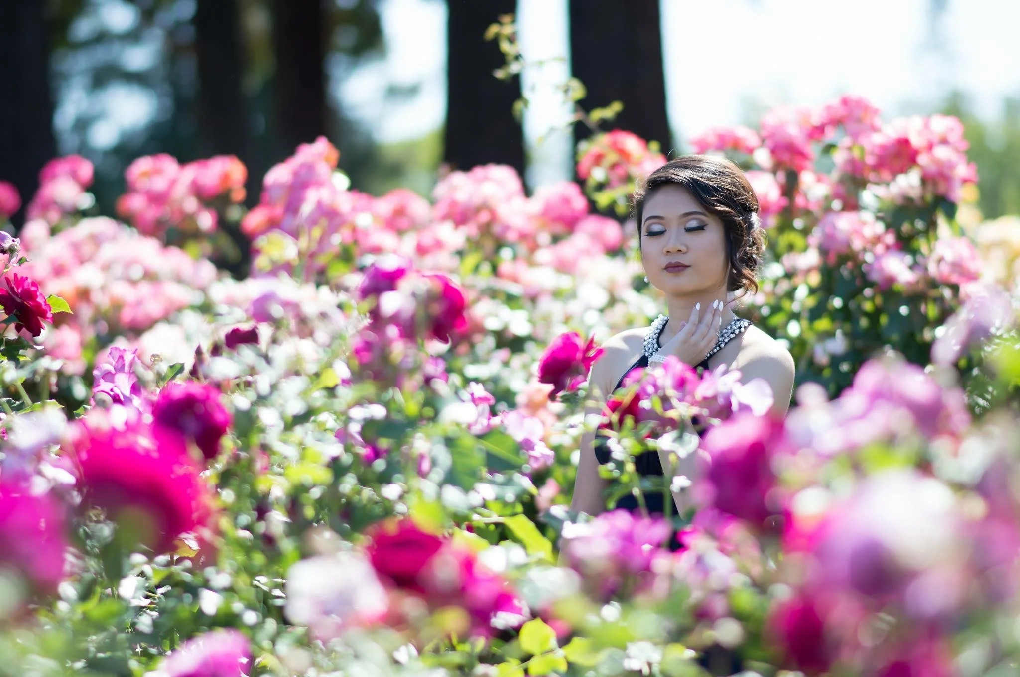 A woman with makeup and jewelry in a dress standing among pink and purple flowers with closed eyes.