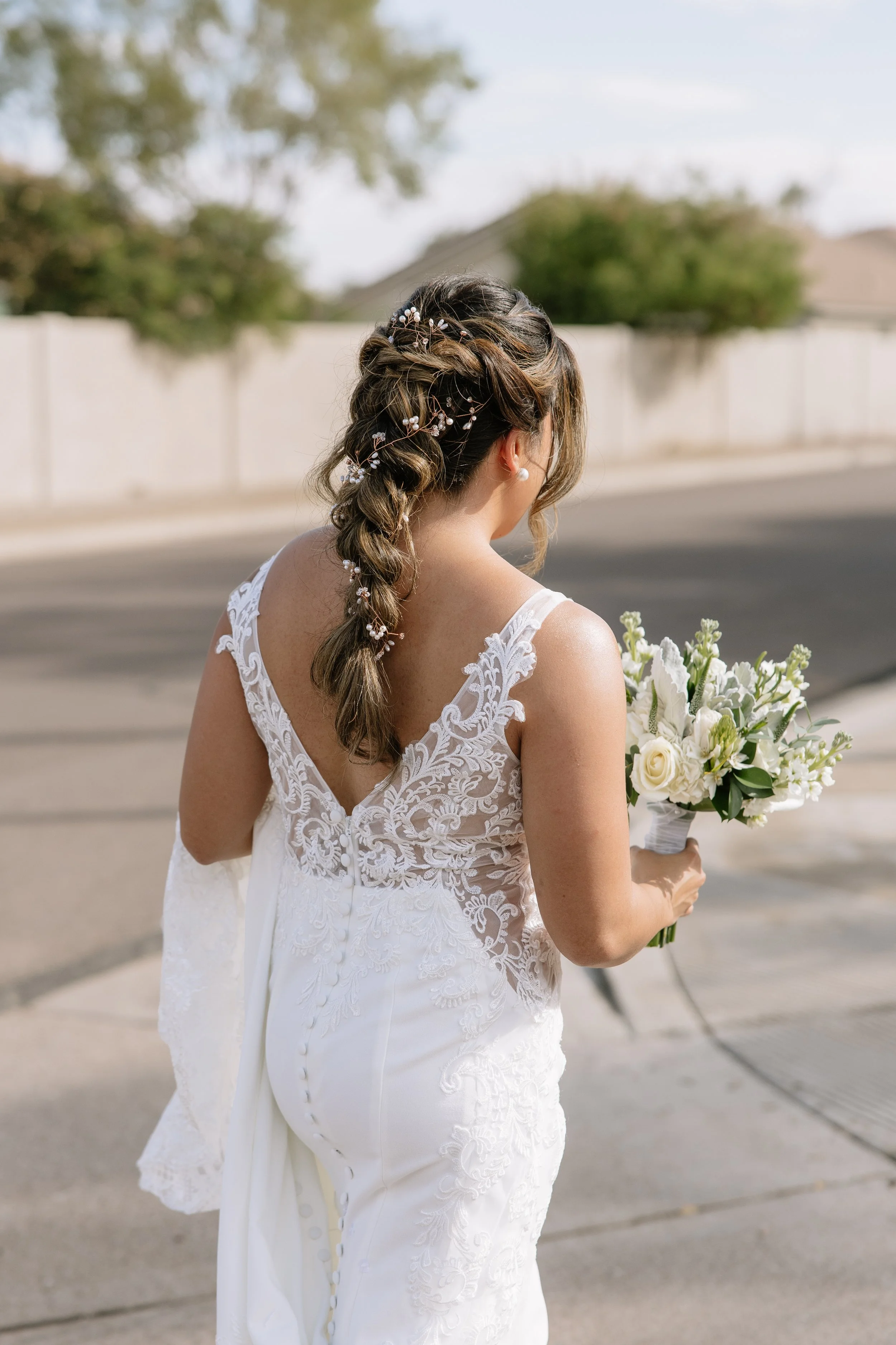 Bridal woman in a white lace wedding dress holding a bouquet of white flowers, with braided hair adorned with small decorative elements, standing on a sidewalk outdoors.