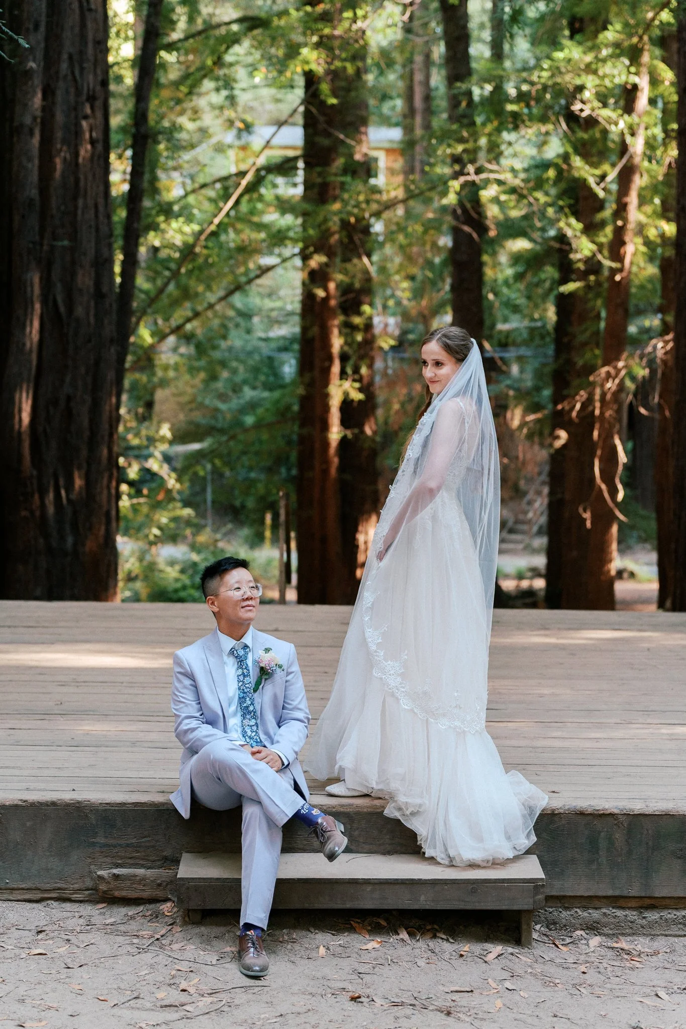 A bride in a white wedding dress and veil standing beside a man in a light gray suit sitting on a wooden platform outdoors, surrounded by tall trees in a forest setting.