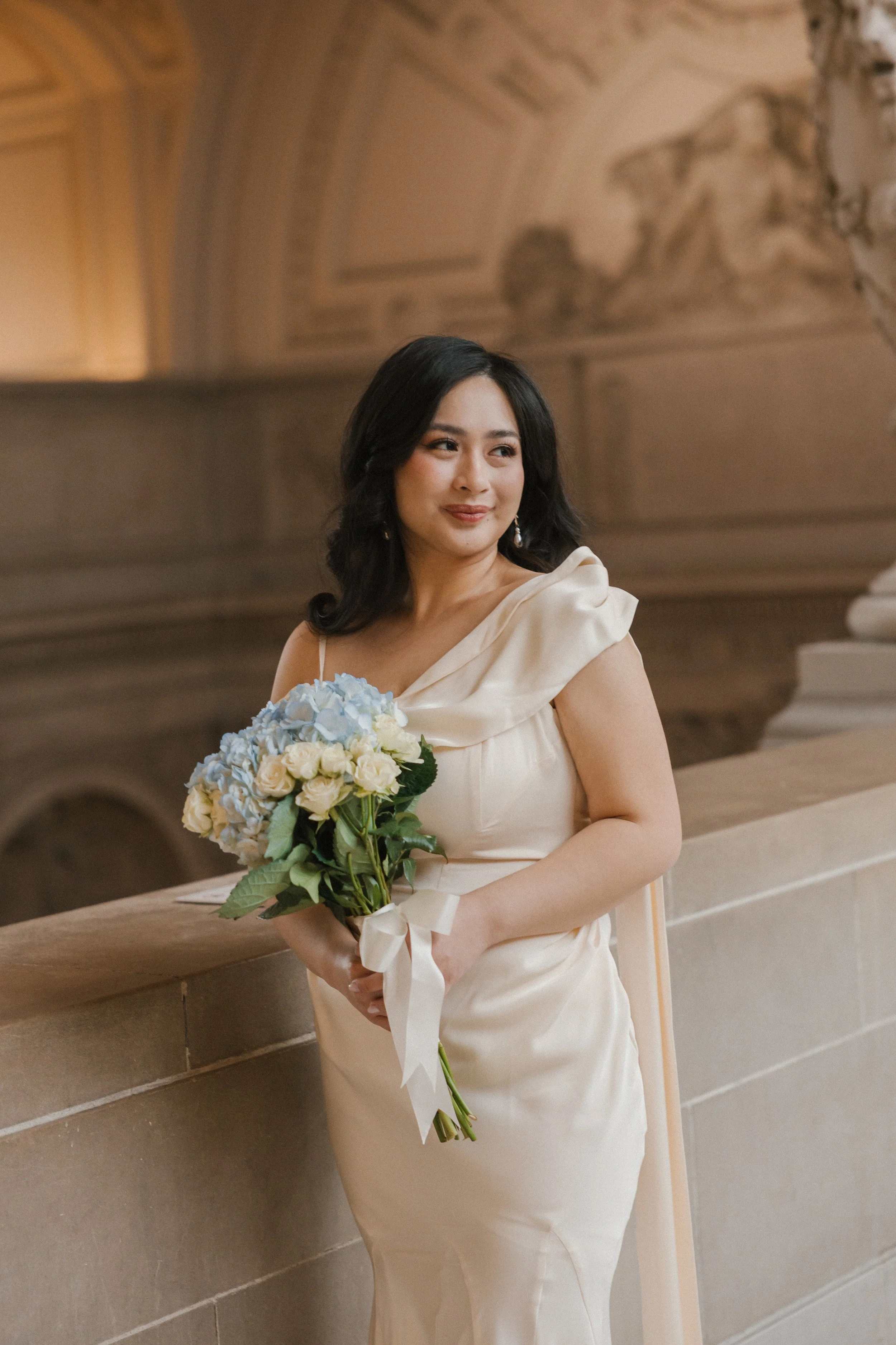 A woman in an elegant cream-colored dress holding a bouquet of white and light blue flowers, standing indoors against a stone railing with ornate architectural details and mural paintings in the background.