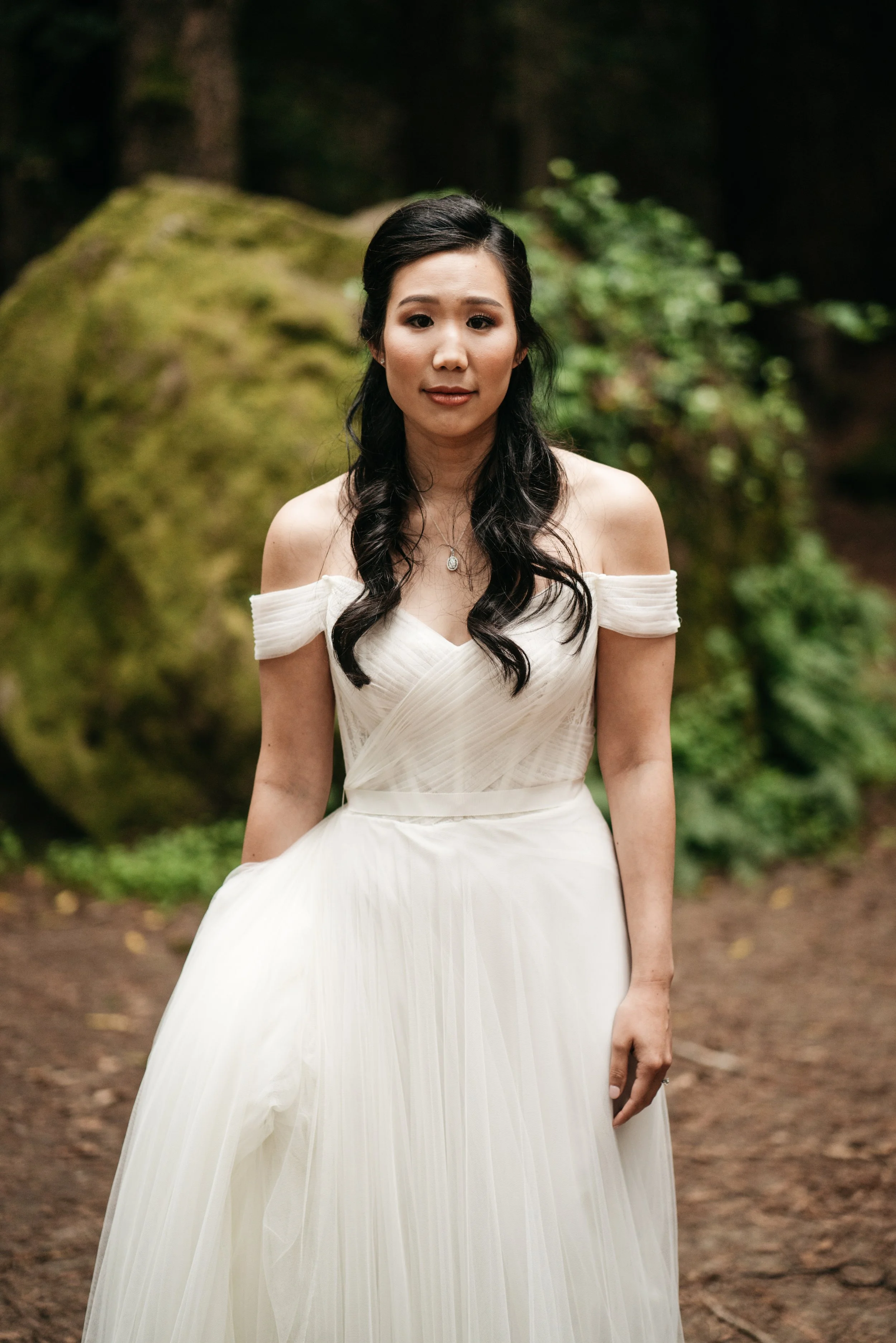 A woman in an off-shoulder white dress standing outdoors in a wooded area.