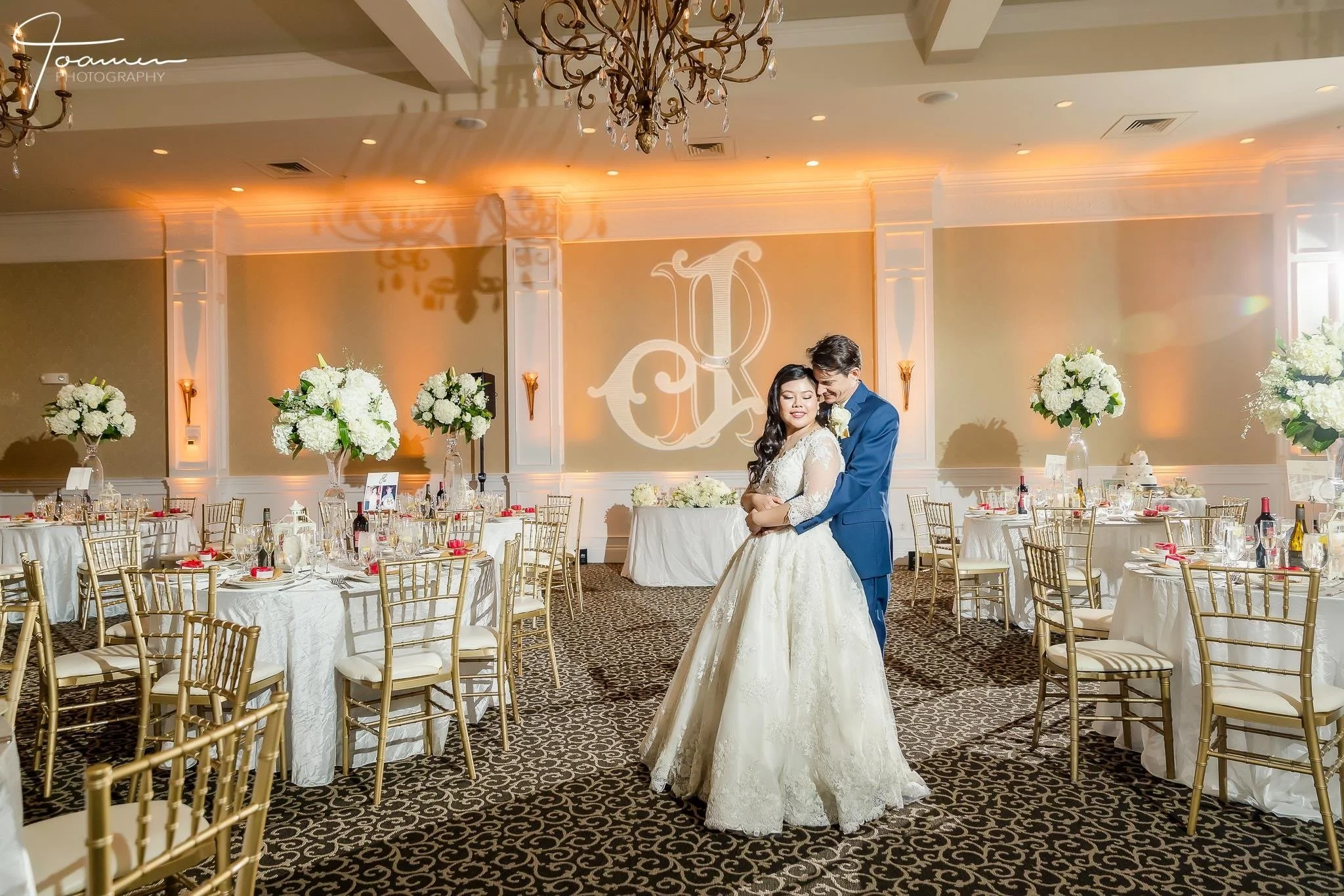 A bride and groom dancing closely at their wedding reception with decorated tables and large floral centerpieces in a banquet hall.