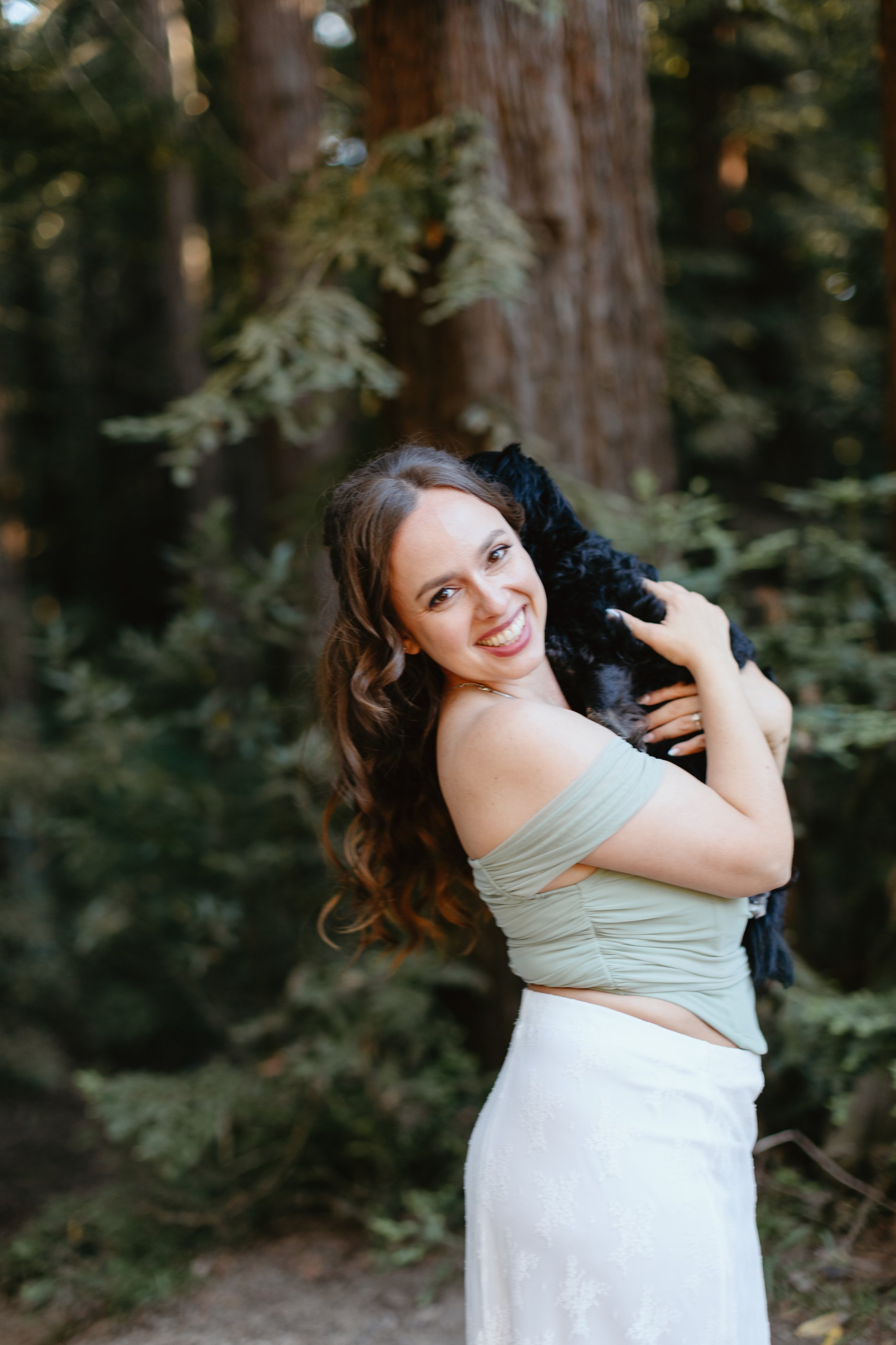 A woman with long wavy hair is smiling and holding a black puppy in her arms outdoors in a forested area with large trees and green foliage.