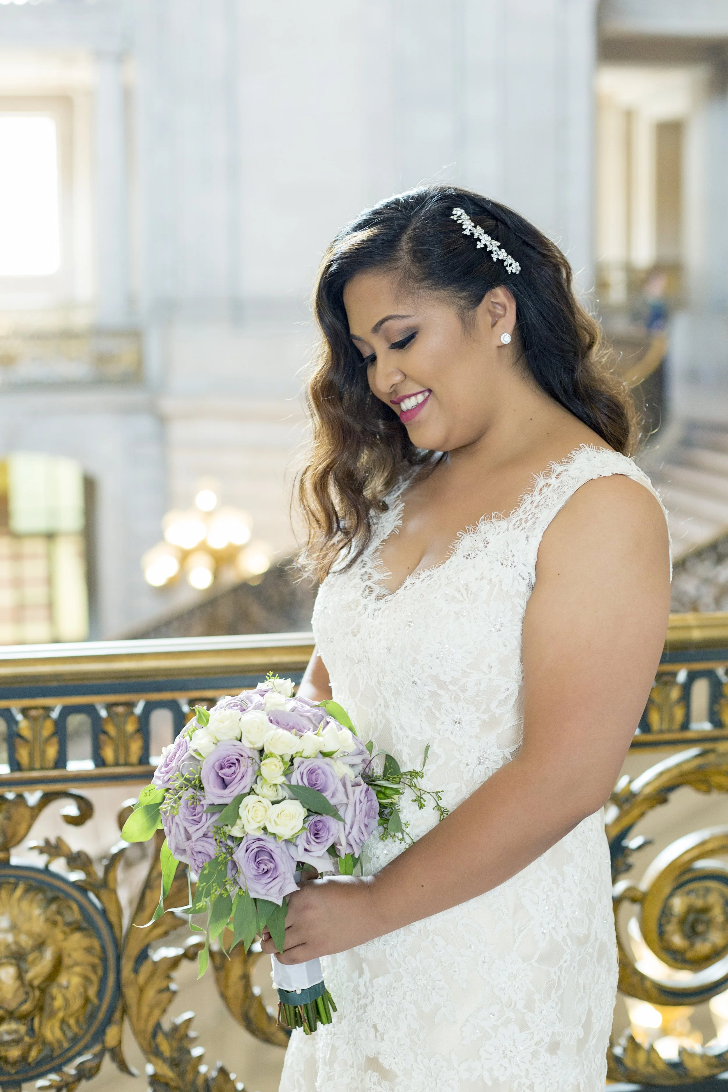 A bride in a white lace wedding dress holding a bouquet of purple and white roses, smiling inside a grand building.